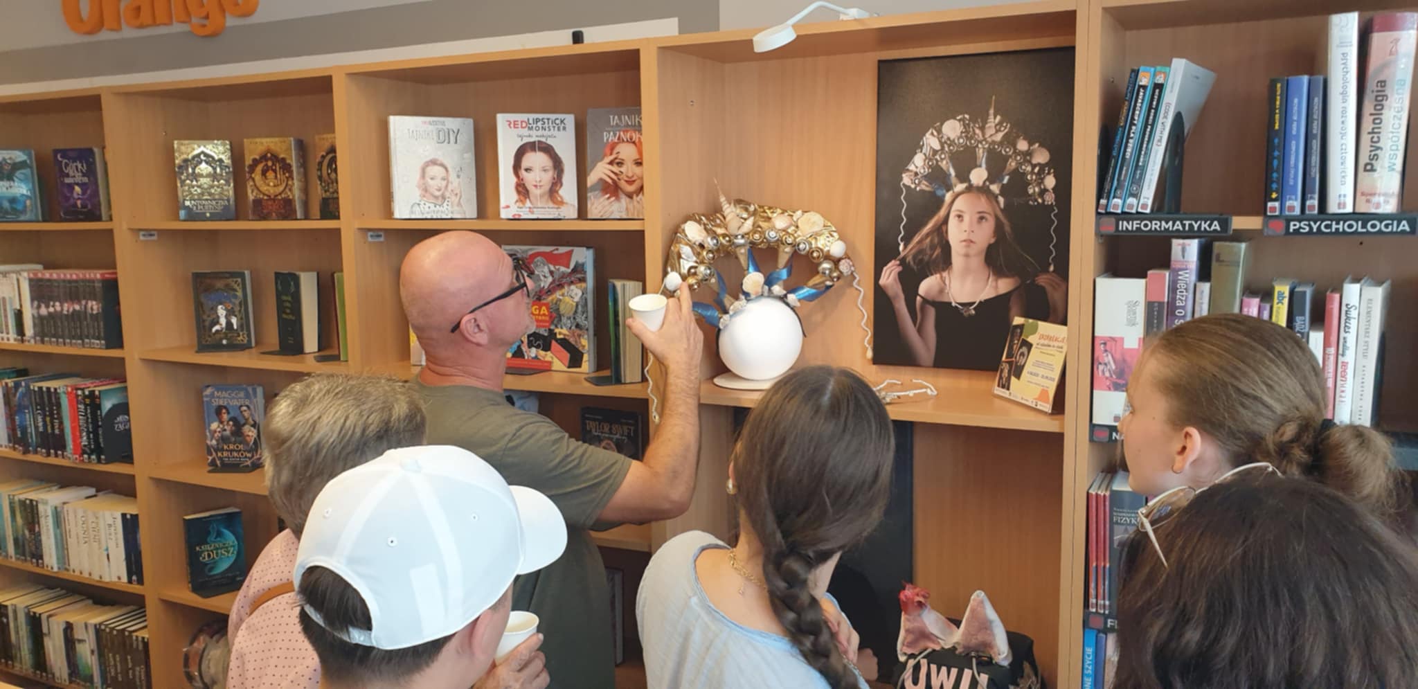 Prezentacja pracy artystycznej na wystawie EkoKreacje – mężczyzna pokazuje szczegóły złoto-niebieskiej korony ozdobionej muszlami, obok fotografia modelki w tym nakryciu głowy, w tle półki z książkami.