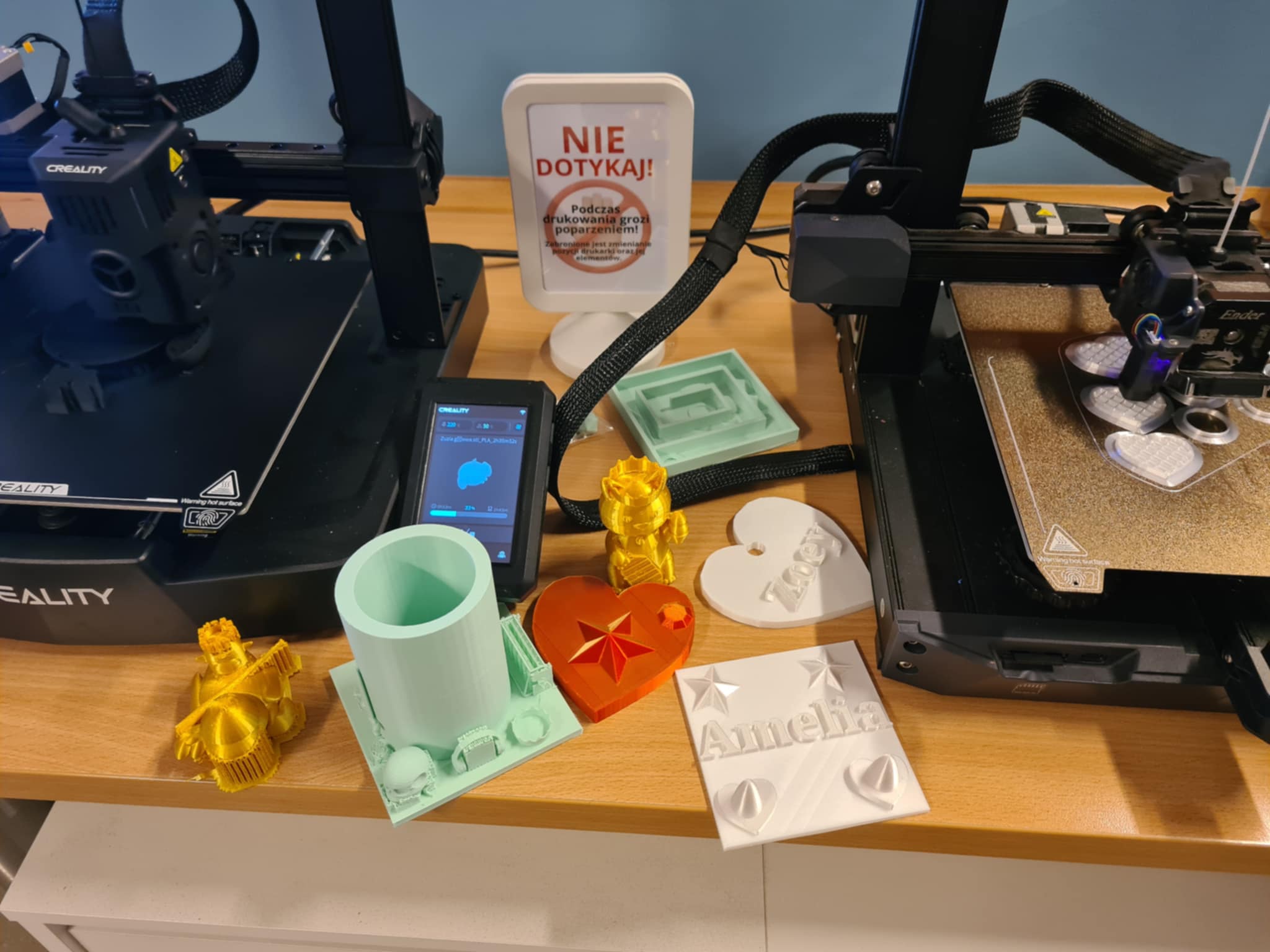 Wydruki 3D w bibliotece – na stole obok drukarek 3D Creality stoją gotowe wydruki: złoty robot, miętowy pojemnik, czerwone serce, białe breloki z imionami „Amelia” i „Zoe”.