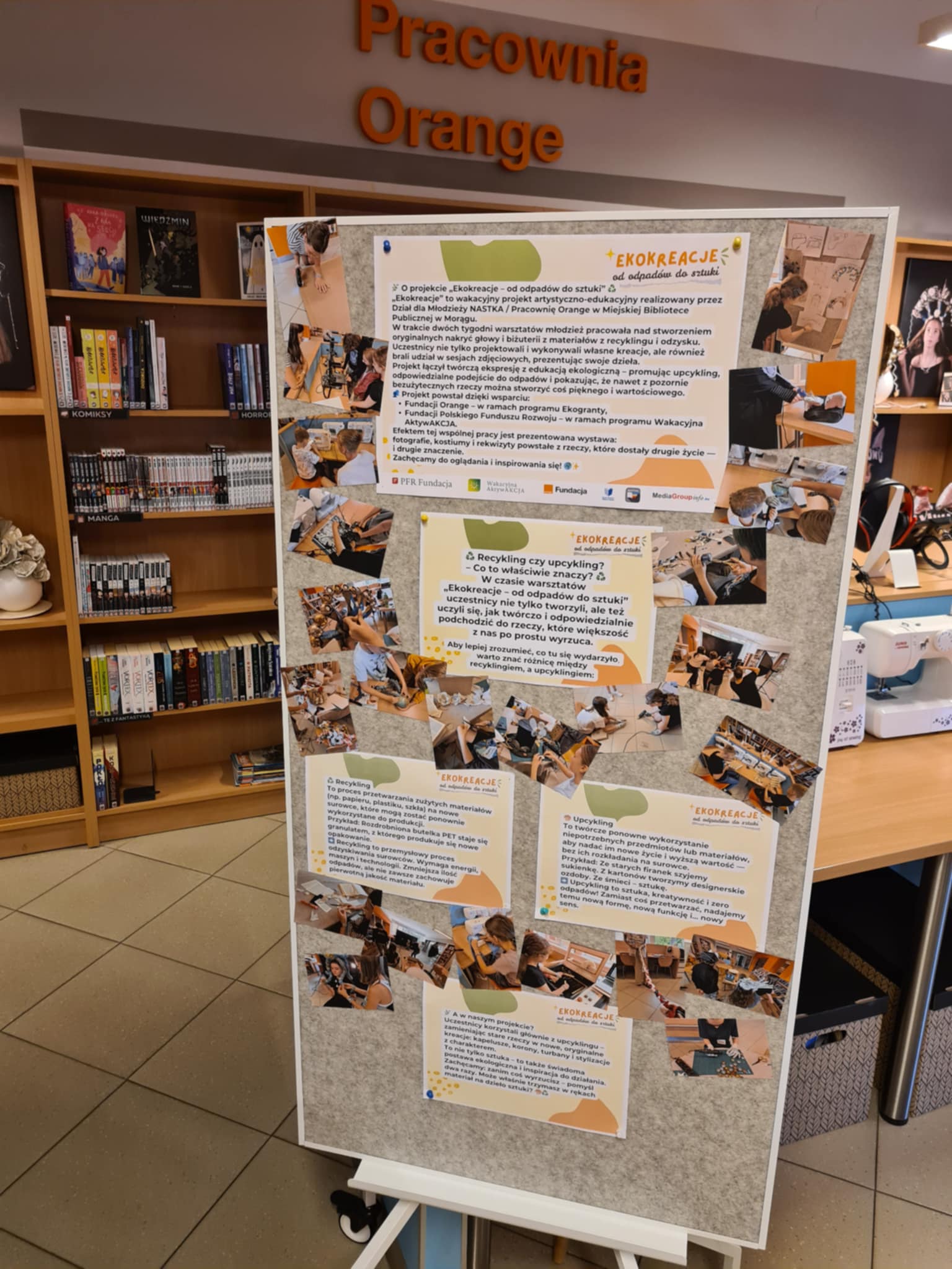 Tablica wystawowa z opisem projektu „Ekokreacje” i zdjęciami z warsztatów, w tle biblioteczne regały.