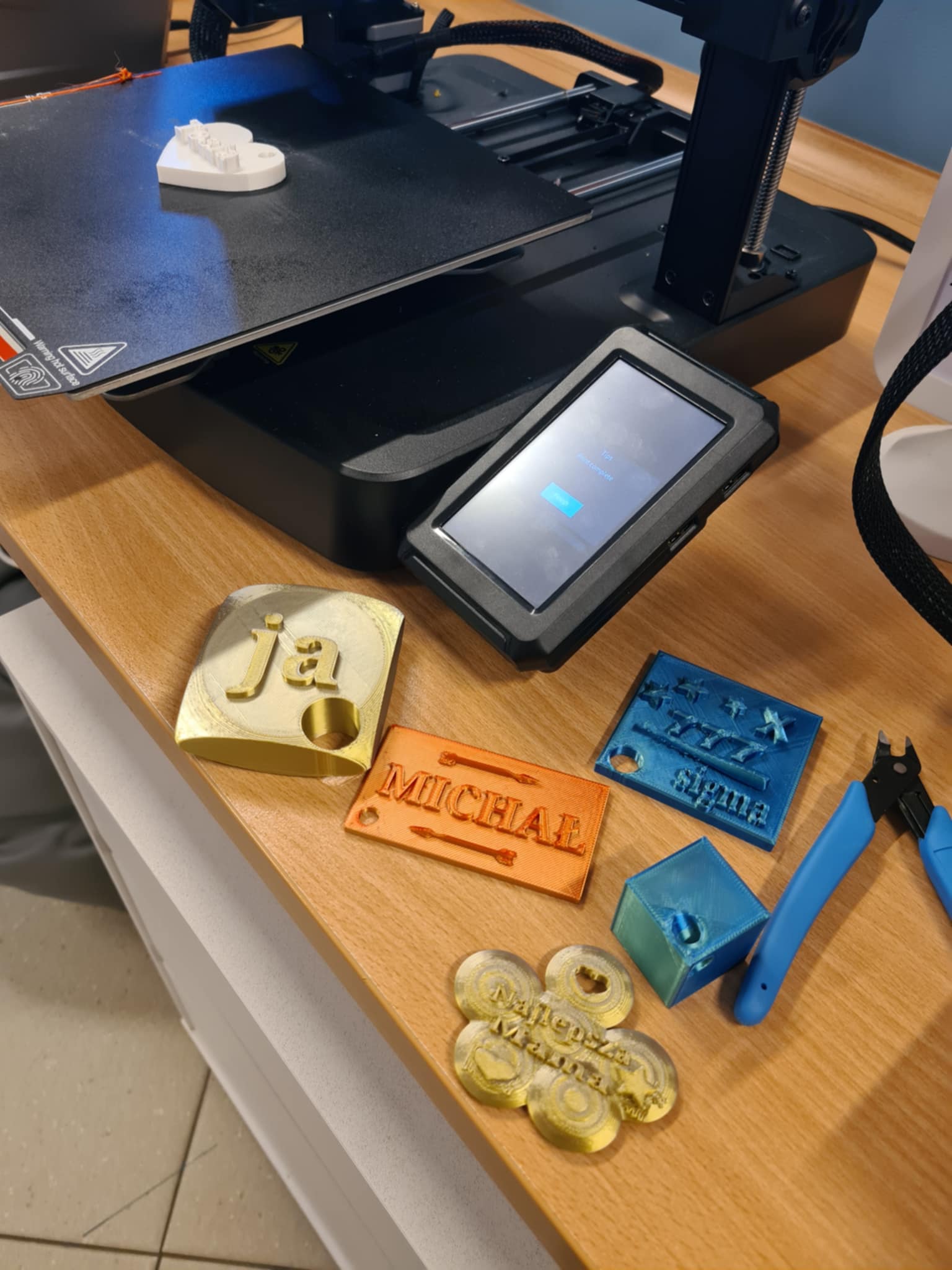 Gotowe wydruki 3D: breloki z imionami i różnymi kształtami, ułożone na stole obok drukarki.