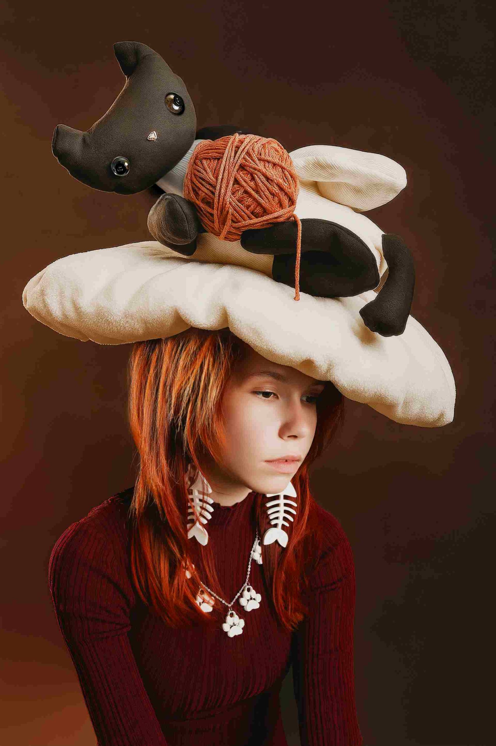 Dziewczyna w bordowym golfie z ogromnym pluszowym kotem i kłębkiem włóczki na kapeluszu, biżuteria w kształcie rybich ości.