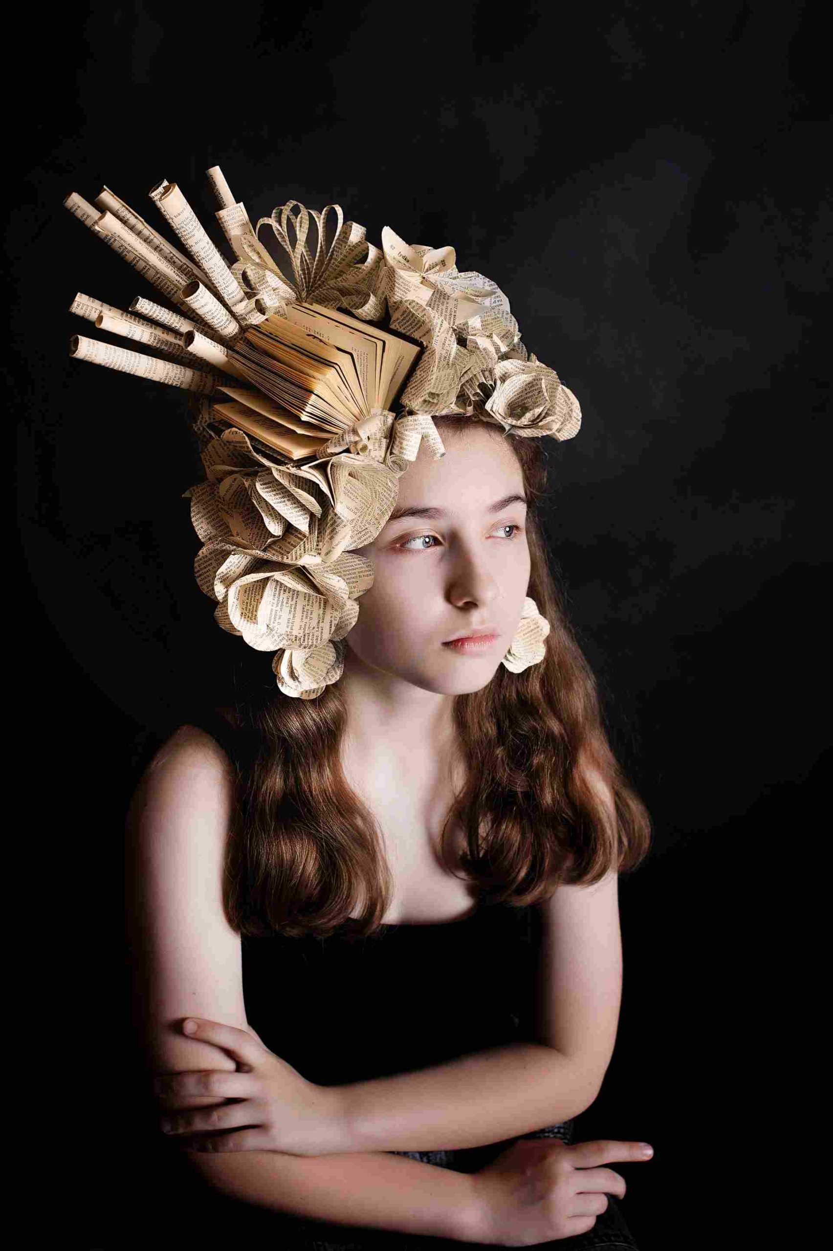 Dziewczyna w spektakularnym kapeluszu z papierowych kwiatów i rozłożonej książki, wykonanym z recyklingu.