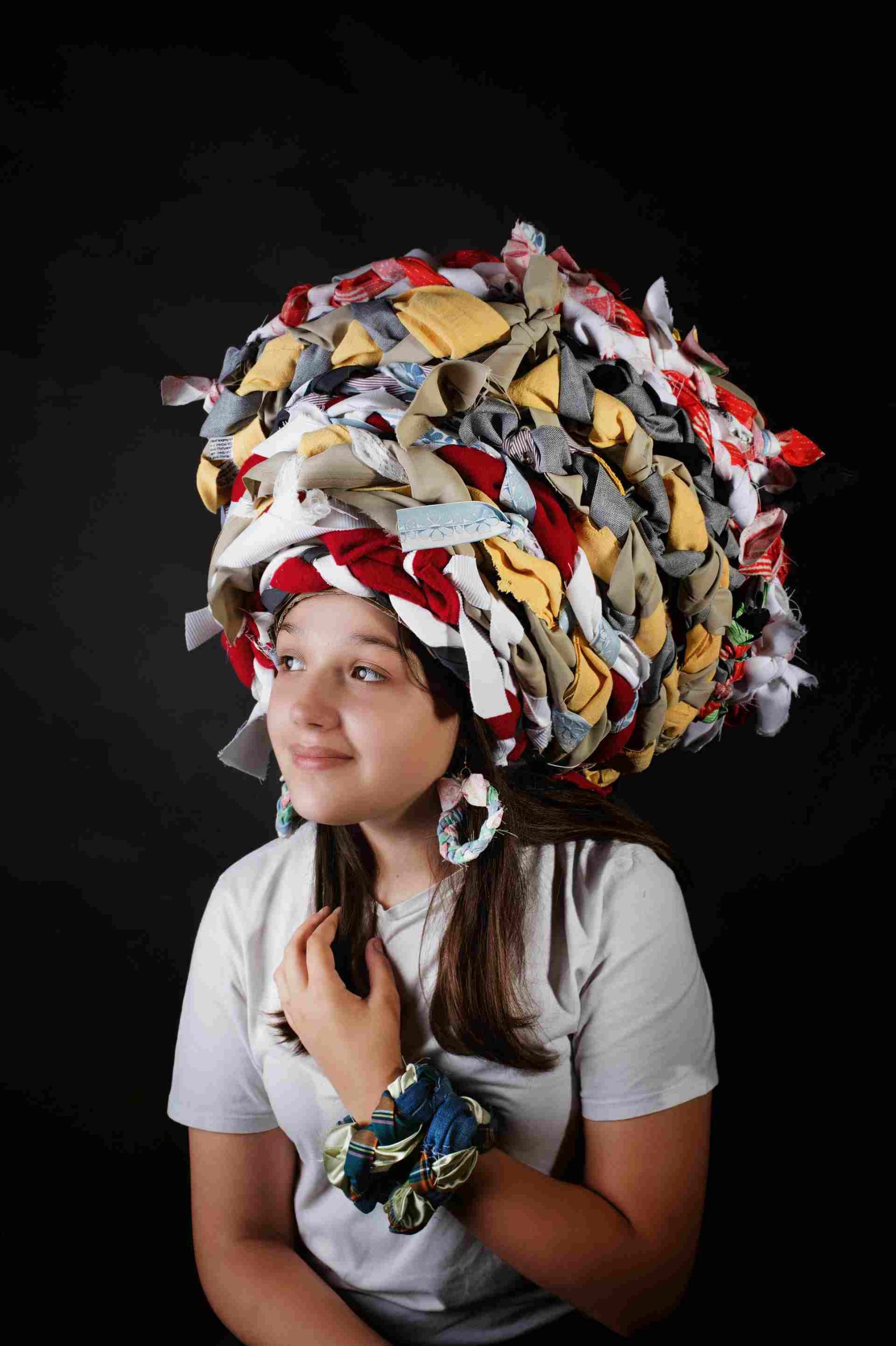 Dziewczyna w kapeluszu zrobionym z kolorowych pasków materiału, uśmiechnięta, w białej koszulce.