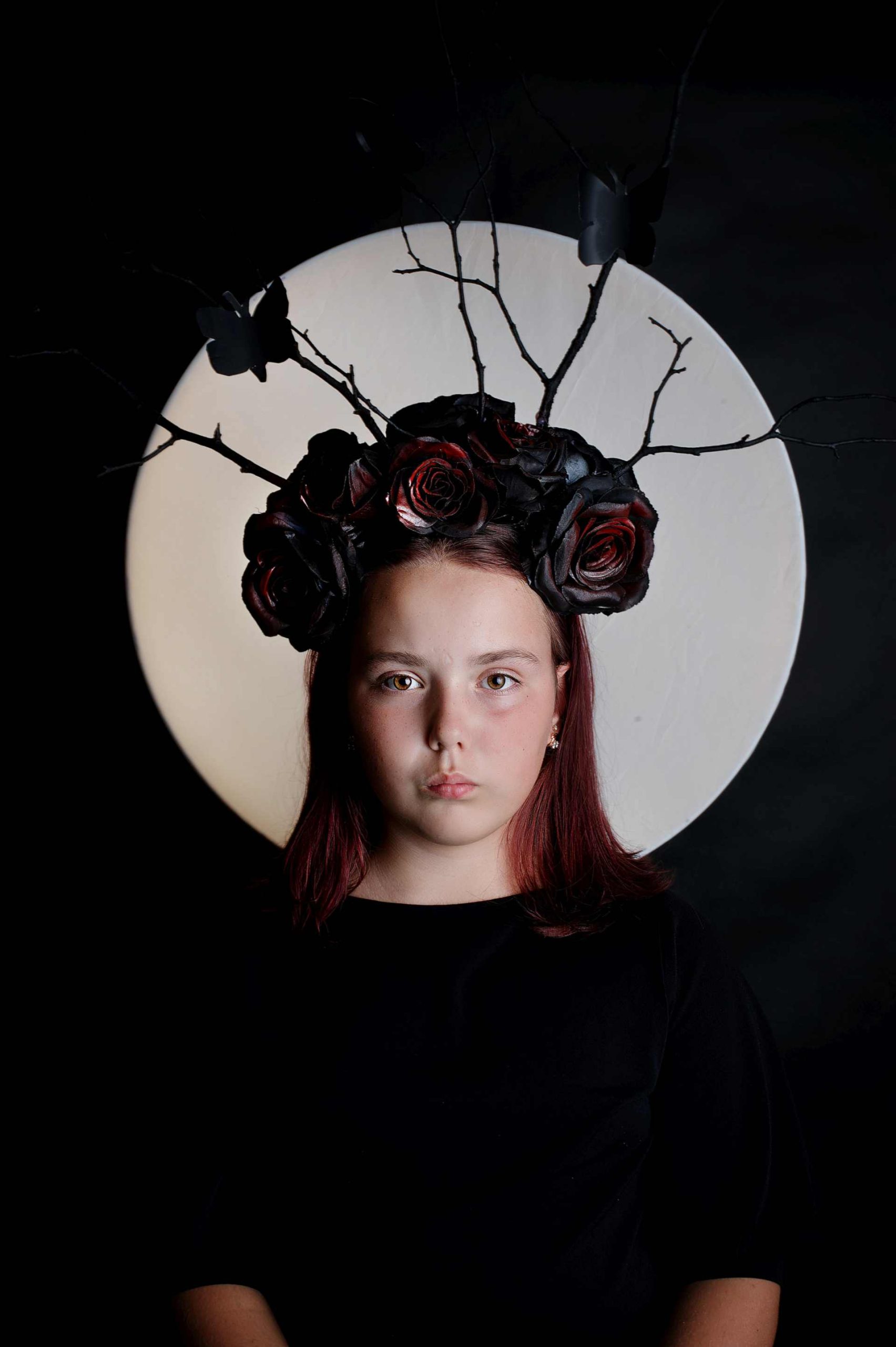Dziewczyna w czarnej sukni, w kapeluszu z czarnych róż, gałązek i motyli, na tle okrągłego światła.