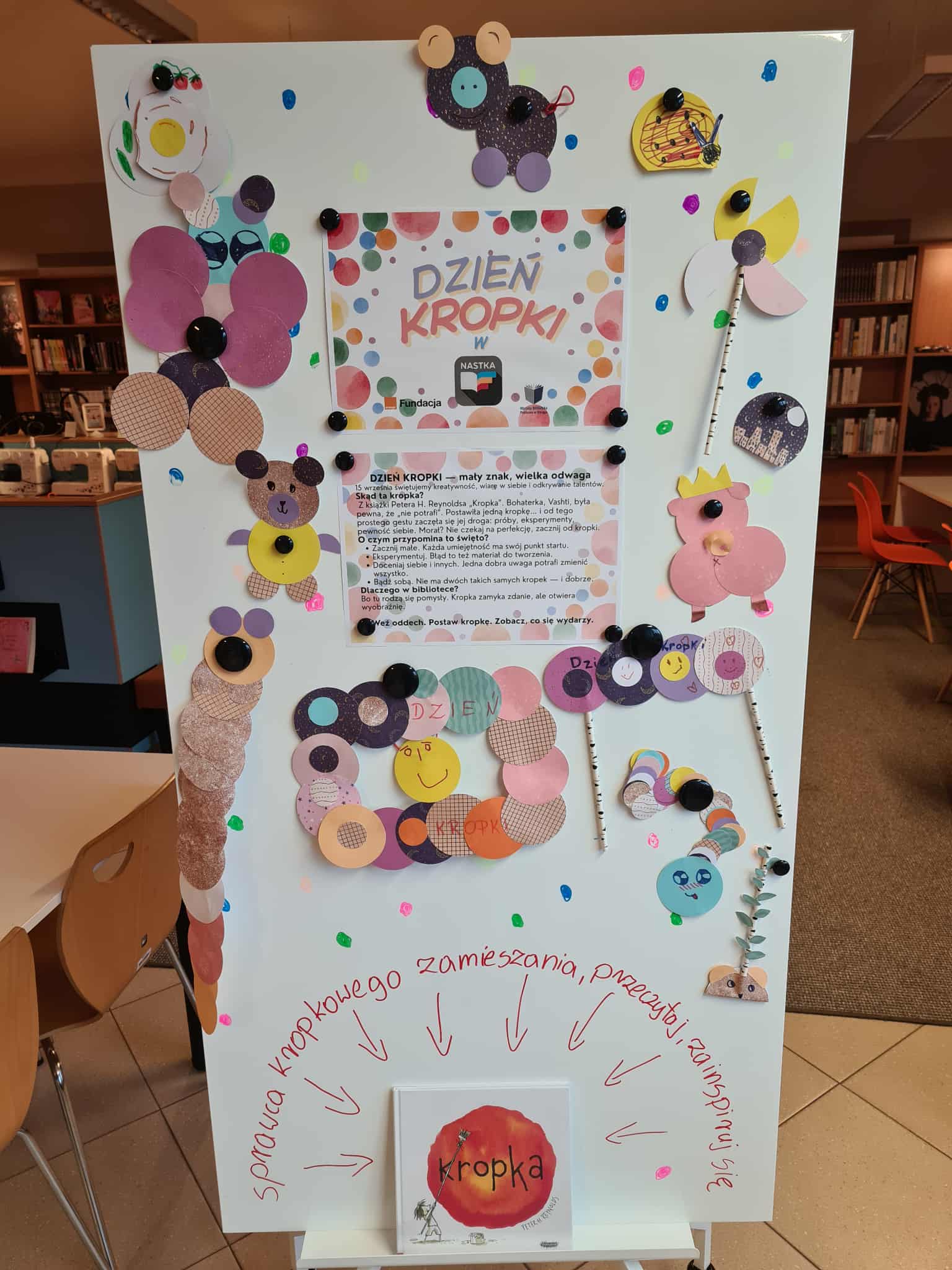 Tablica ozdobiona kolorowymi kropkami i pracami plastycznymi dzieci: papierowe misie, motyle i inne kształty. W centrum plakat „Dzień Kropki w NASTCE”.