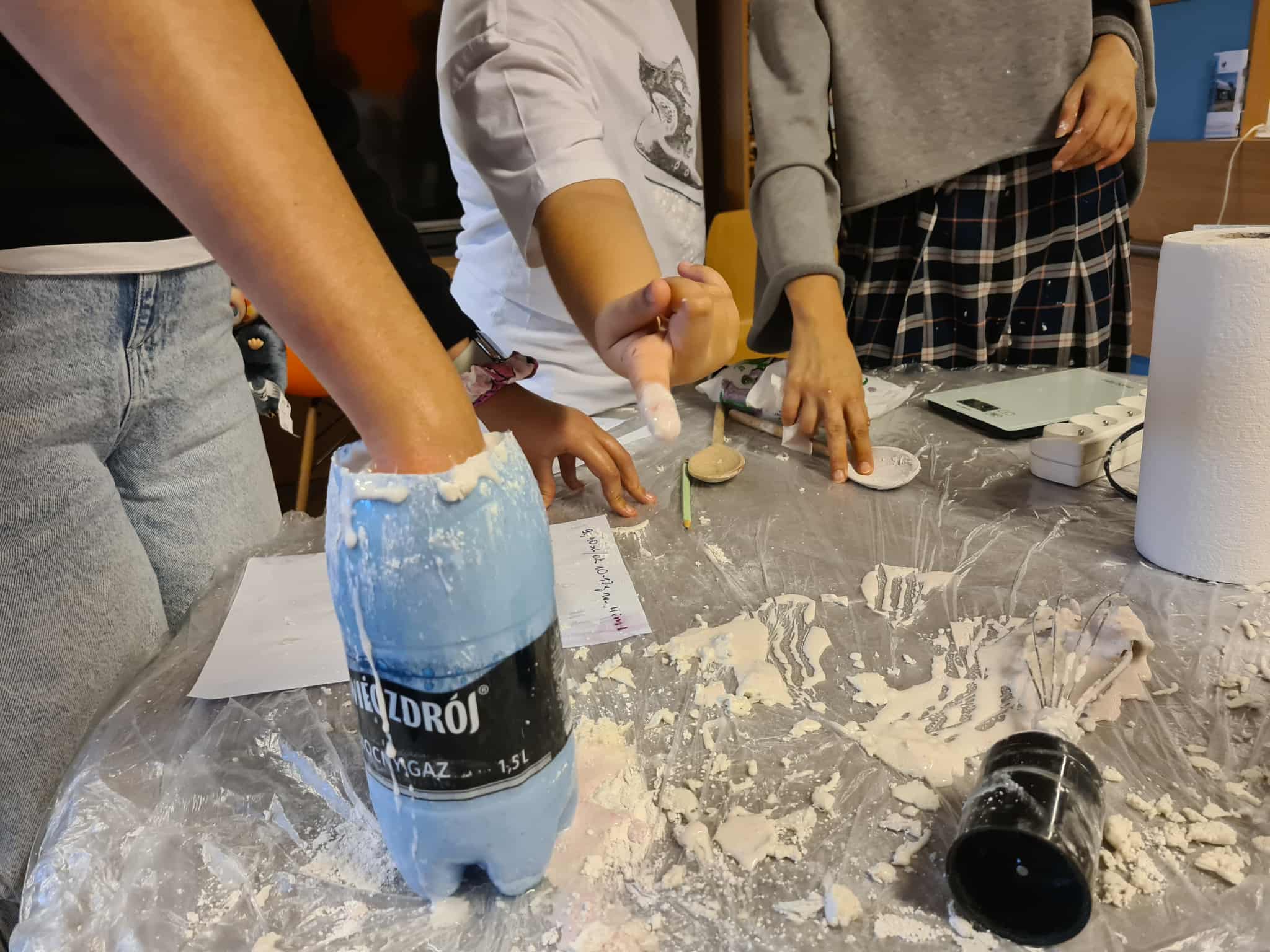 Ręka zanurzona w niebieskiej butelce plastikowej wypełnionej masą do odlewów, na stole widać bałagan po pracy.