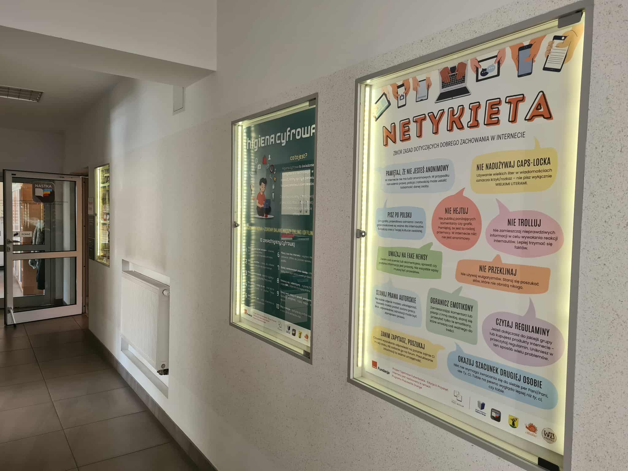 Korytarz biblioteki z widocznymi podświetlanymi gablotami. W gablotach plakaty: po lewej o higienie cyfrowej, po prawej o zasadach netykiety. Na drzwiach w tle widnieje logo działu dla młodzieży NASTKA.