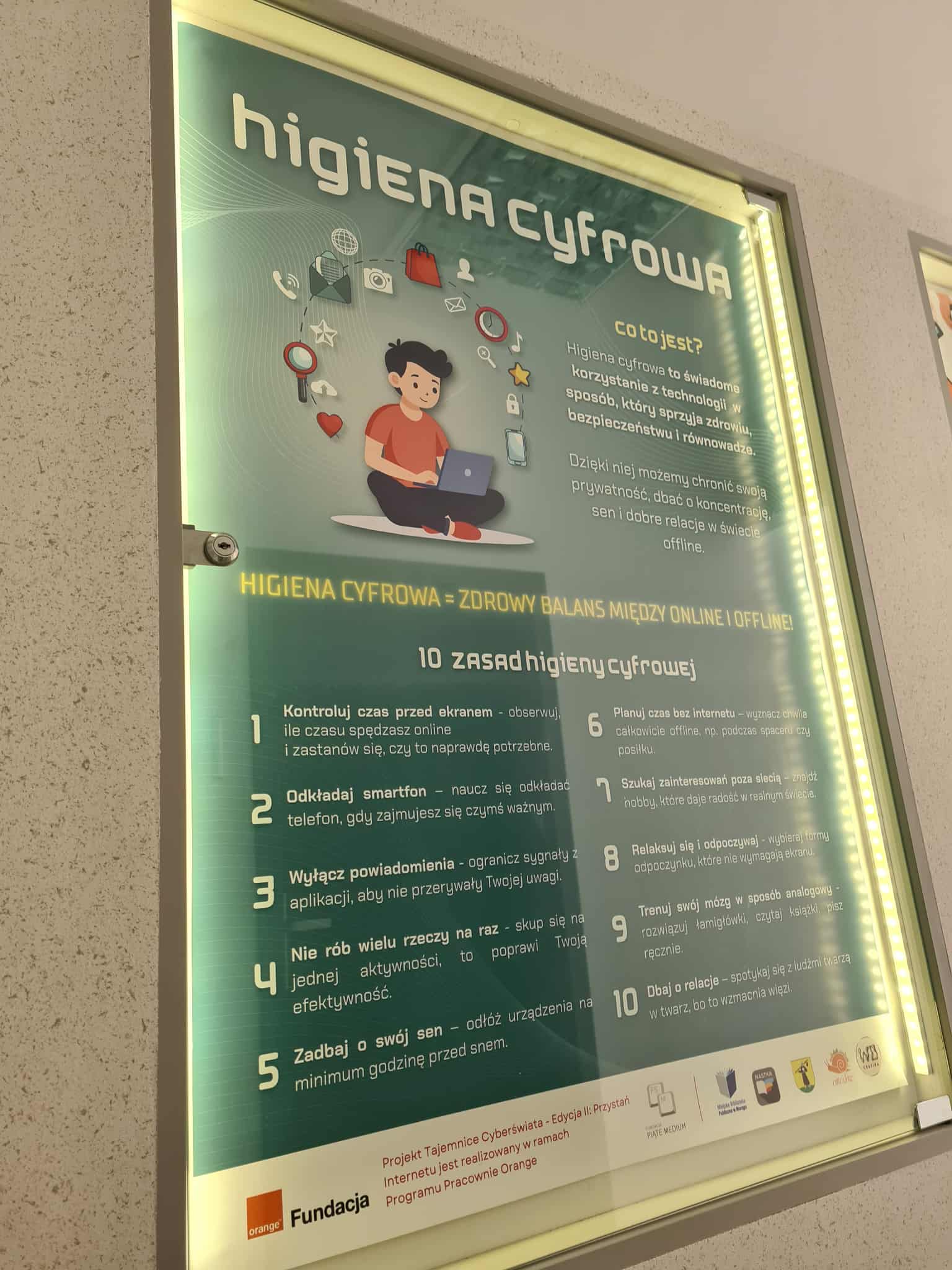 Plakat o higienie cyfrowej w gablocie. Zawiera ilustrację postaci z laptopem oraz wypisane zasady świadomego korzystania z technologii, np. kontrola czasu przed ekranem, odkładanie smartfona, wyłączanie powiadomień.
