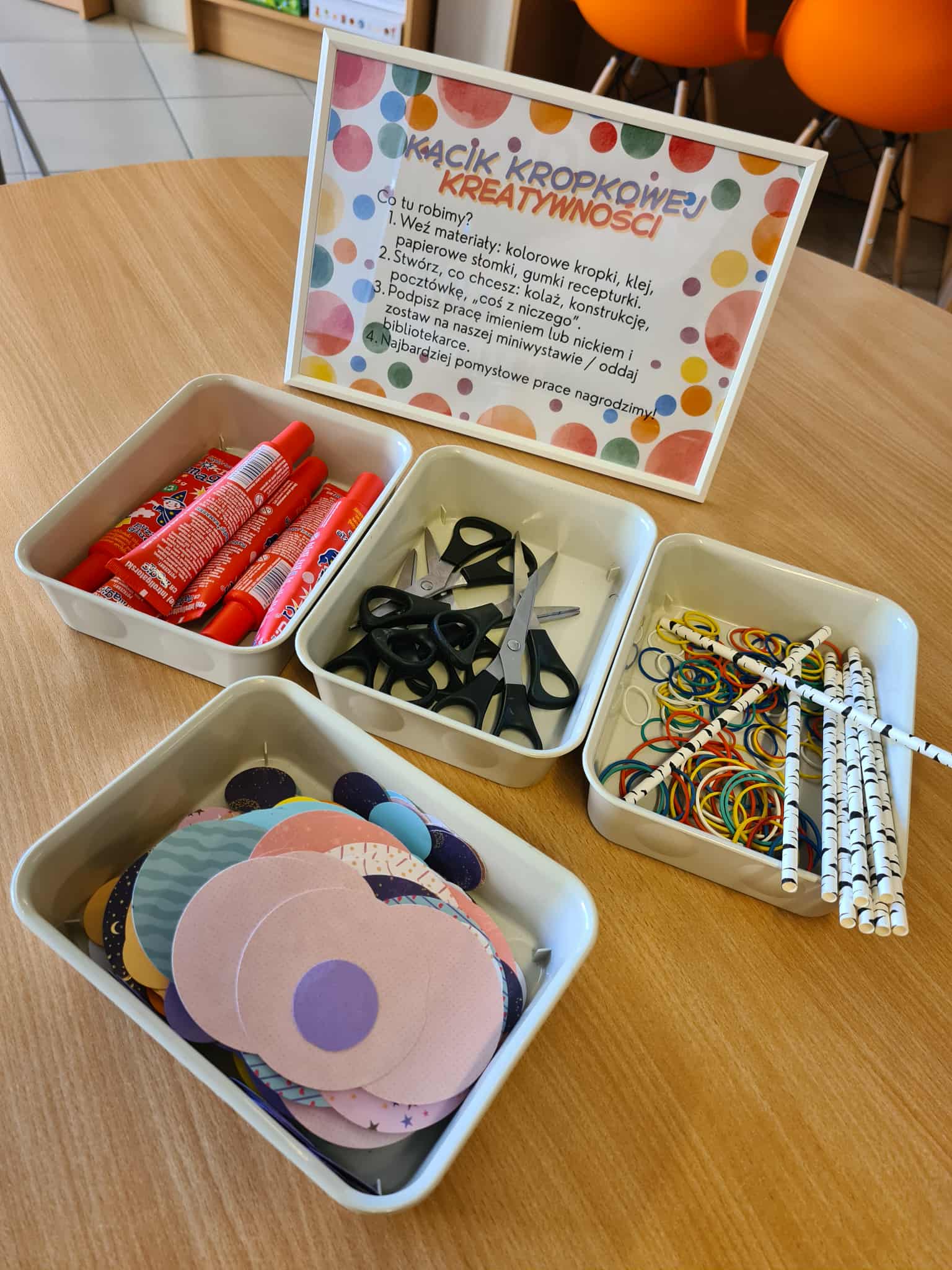 Pojemniki z przyborami: kolorowe kółka papierowe, kleje, nożyczki, słomki i gumki recepturki, przygotowane do pracy warsztatowej.