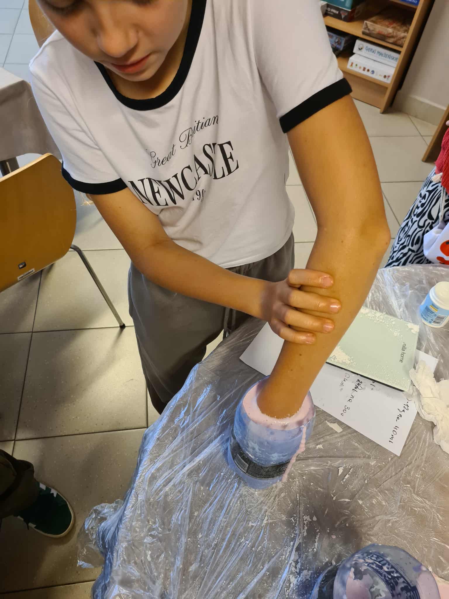 Dziewczynka zanurza rękę w różowej masie w plastikowej butelce, stojąc przy stole zabezpieczonym folią.