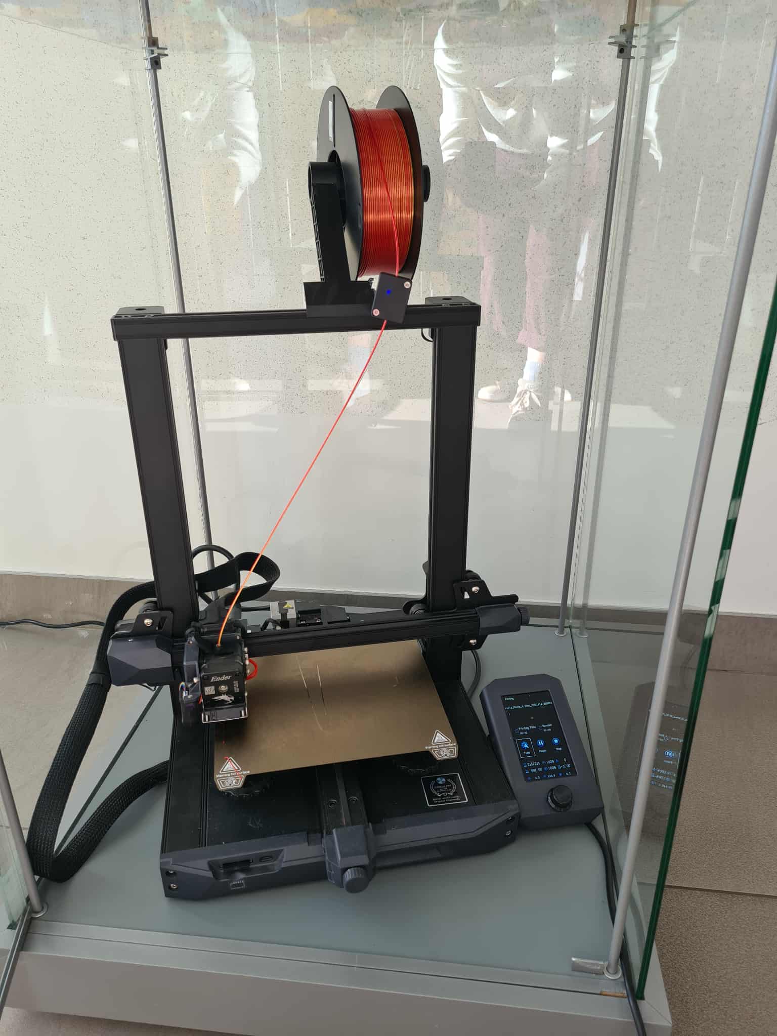 Drukarka 3D Ender stojąca w szklanej gablocie, przygotowana do pracy z pomarańczowym filamentem.