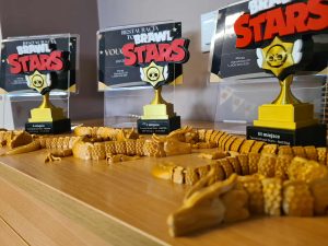 rzy statuetki z napisem Brawl Stars ustawione na blacie bibliotecznym, obok złotych wydruków 3D w kształcie smoków — nagrody dla zwycięzców turnieju.