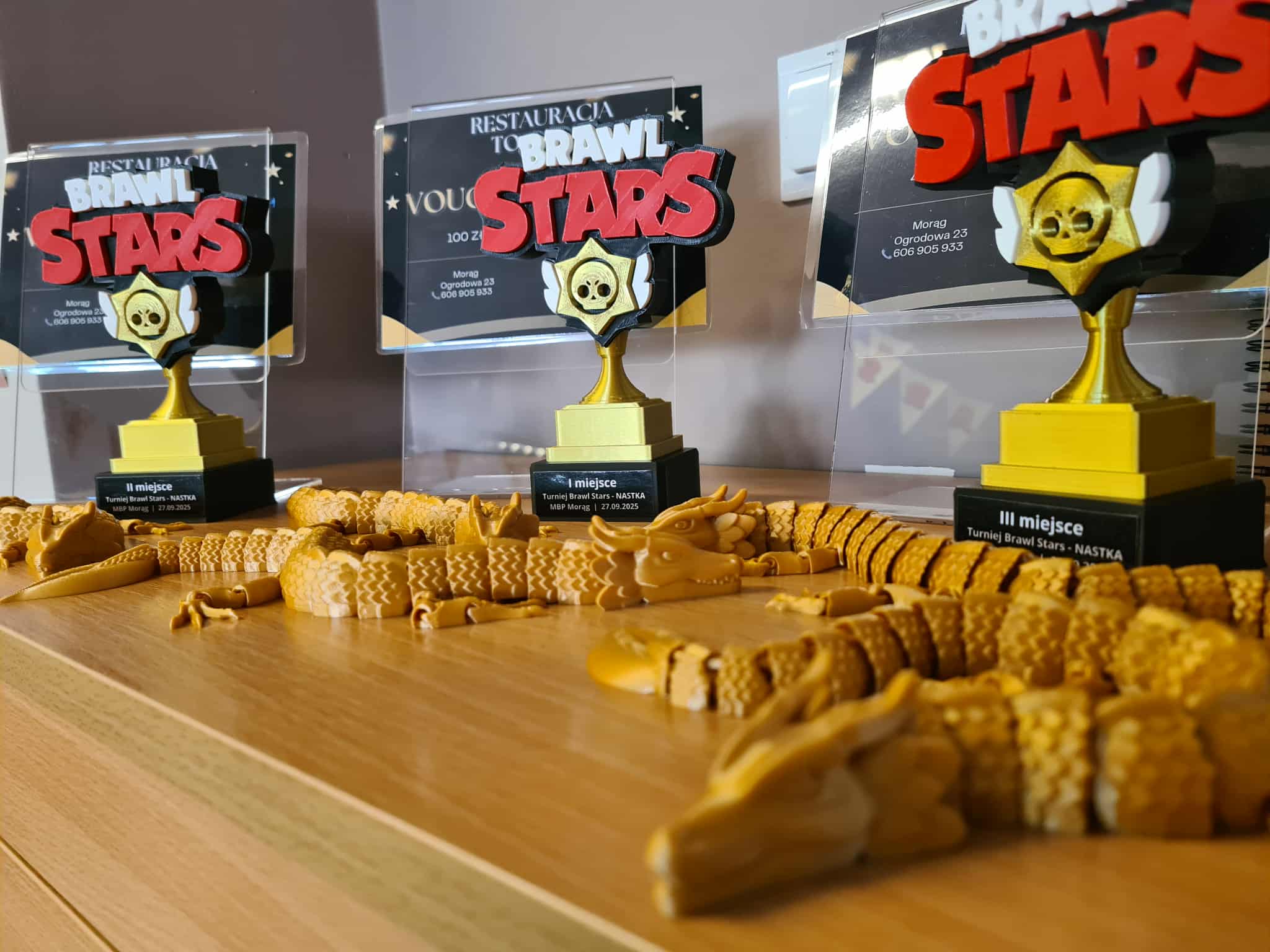 rzy statuetki z napisem Brawl Stars ustawione na blacie bibliotecznym, obok złotych wydruków 3D w kształcie smoków — nagrody dla zwycięzców turnieju.
