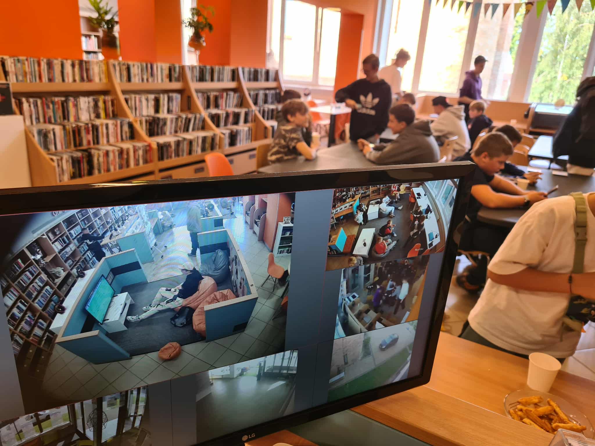 Monitor z podglądem kamer w bibliotece pokazujący graczy przy stolikach i w strefie gier, w tle młodzież grająca na telefonach w kolorowej sali działu NASTKA.