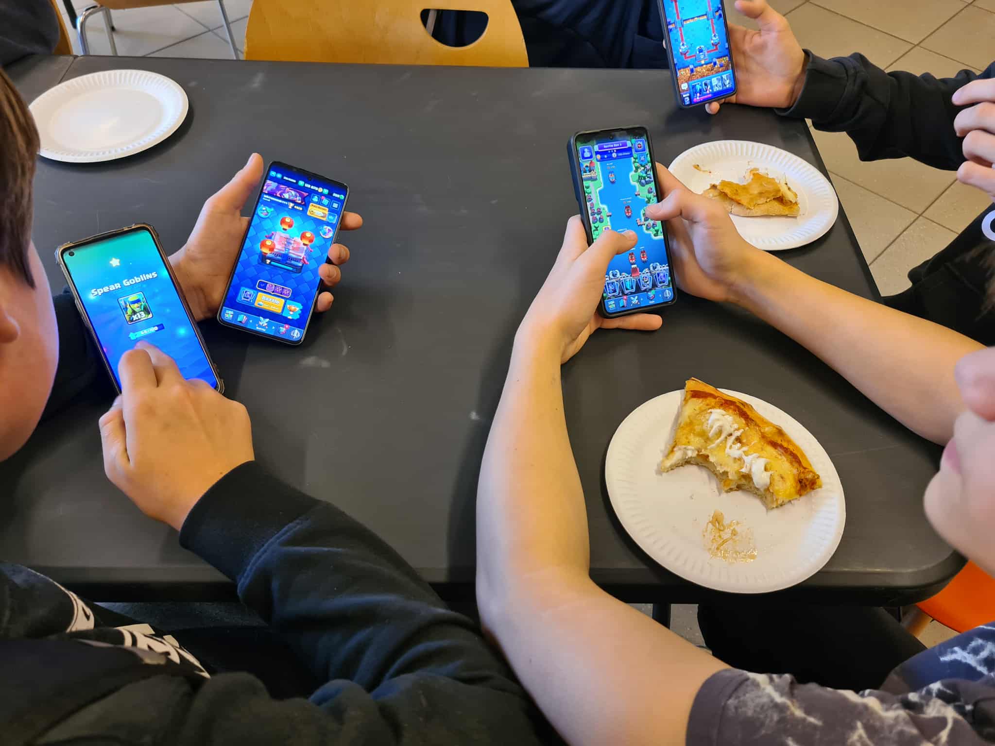 Zbliżenie na dłonie kilku uczestników trzymających smartfony z włączoną grą Brawl Stars. Na stole papierowe talerzyki z kawałkami pizzy.