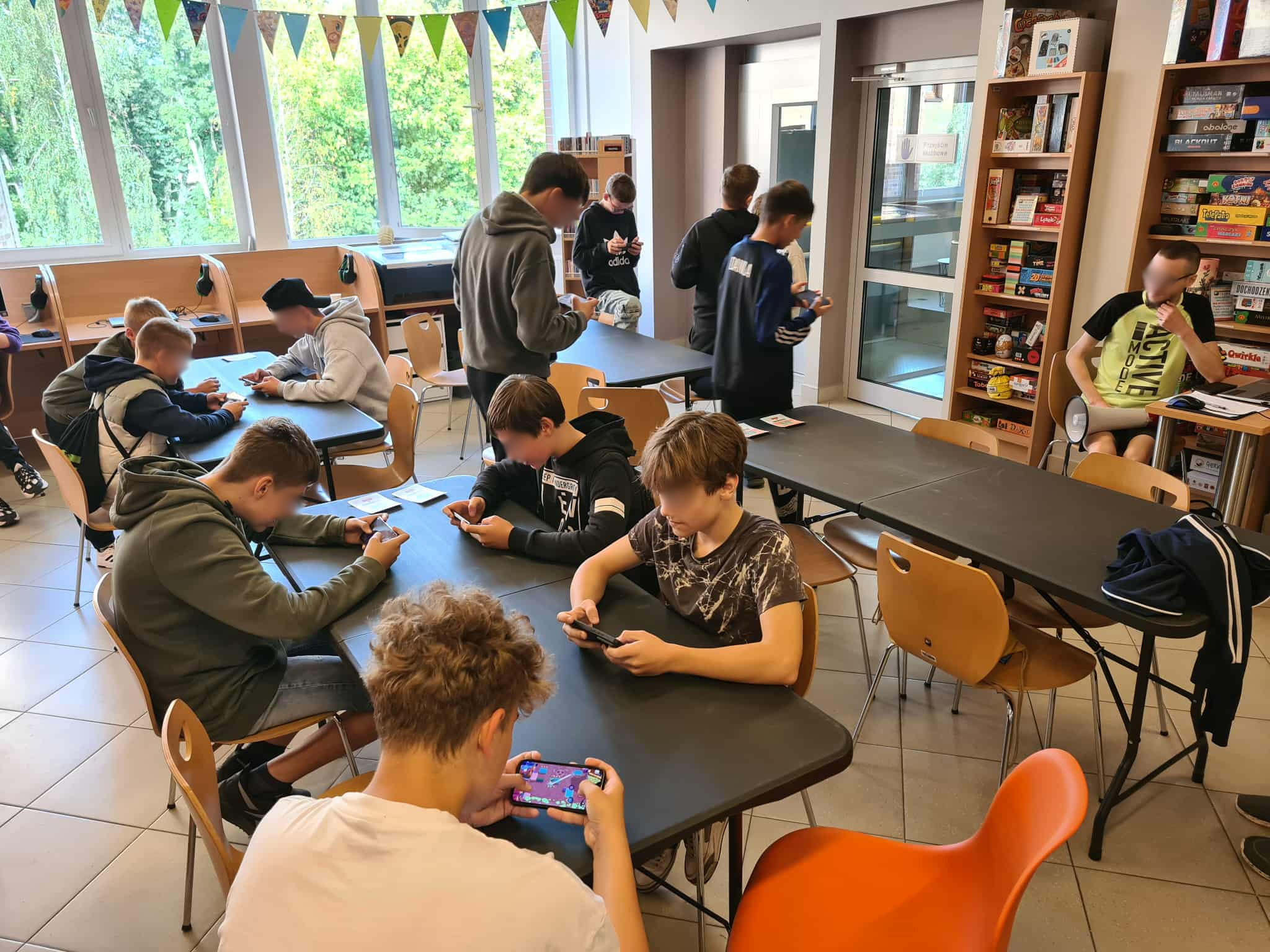 Uczestnicy turnieju siedzą przy stołach w bibliotece i grają w Brawl Stars na swoich telefonach. W tle regały z książkami i dekoracje w kolorowe chorągiewki.