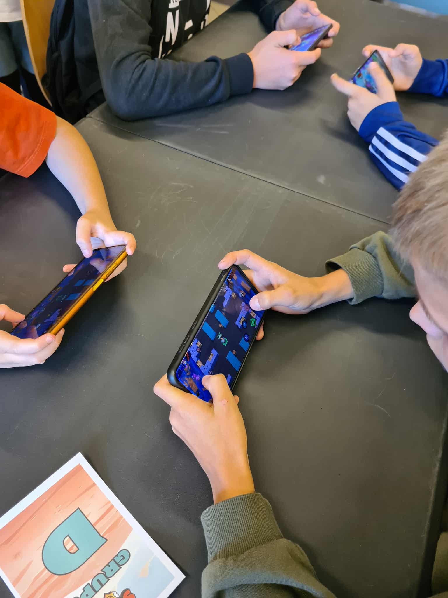 Zbliżenie na dłonie graczy trzymających telefony z uruchomioną grą Brawl Stars. Na stole widoczna kartka z oznaczeniem grupy „D”.