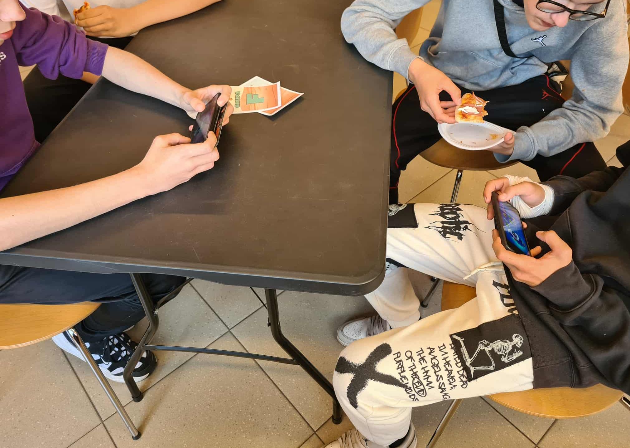 rzej gracze siedzą przy stole z telefonami w dłoniach, grają w Brawl Stars. Jeden z uczestników je kawałek pizzy z papierowego talerzyka.