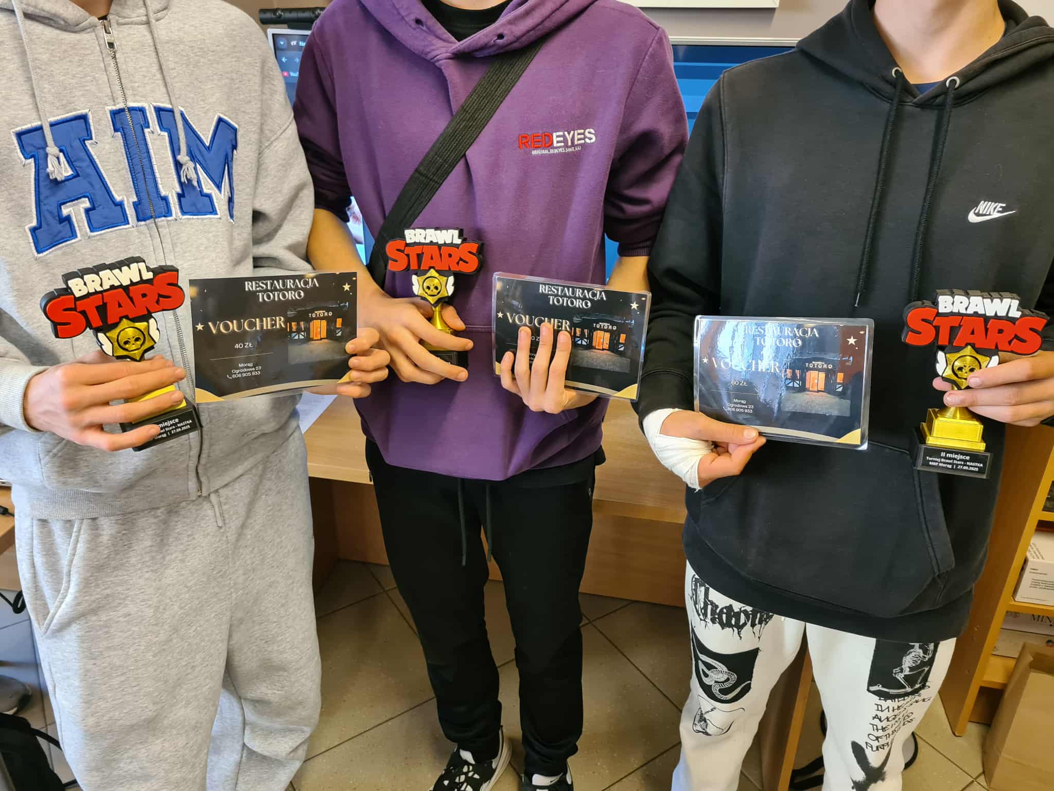 Trzech chłopców trzyma w dłoniach puchary z logo gry Brawl Stars i vouchery do restauracji Totoro — nagrody za udział w turnieju w bibliotece.