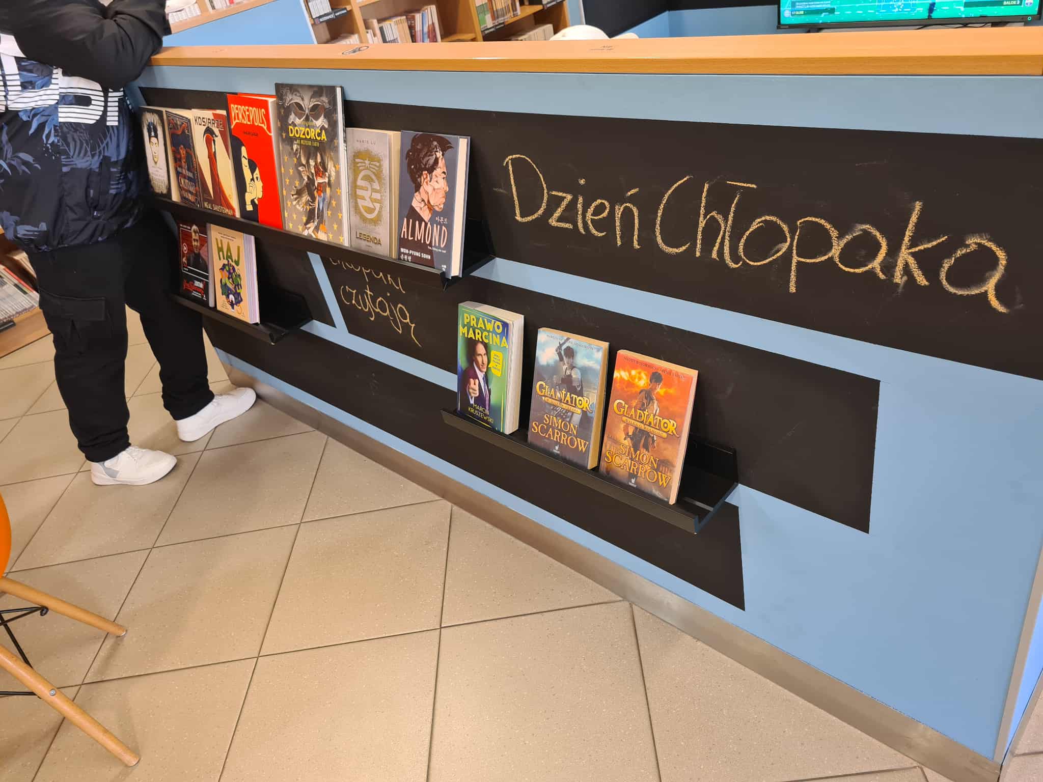 Fragment bibliotecznej lady z książkami wystawionymi na półce oraz napisem „Dzień Chłopaka” zapisanym kredą na czarnej tablicy.