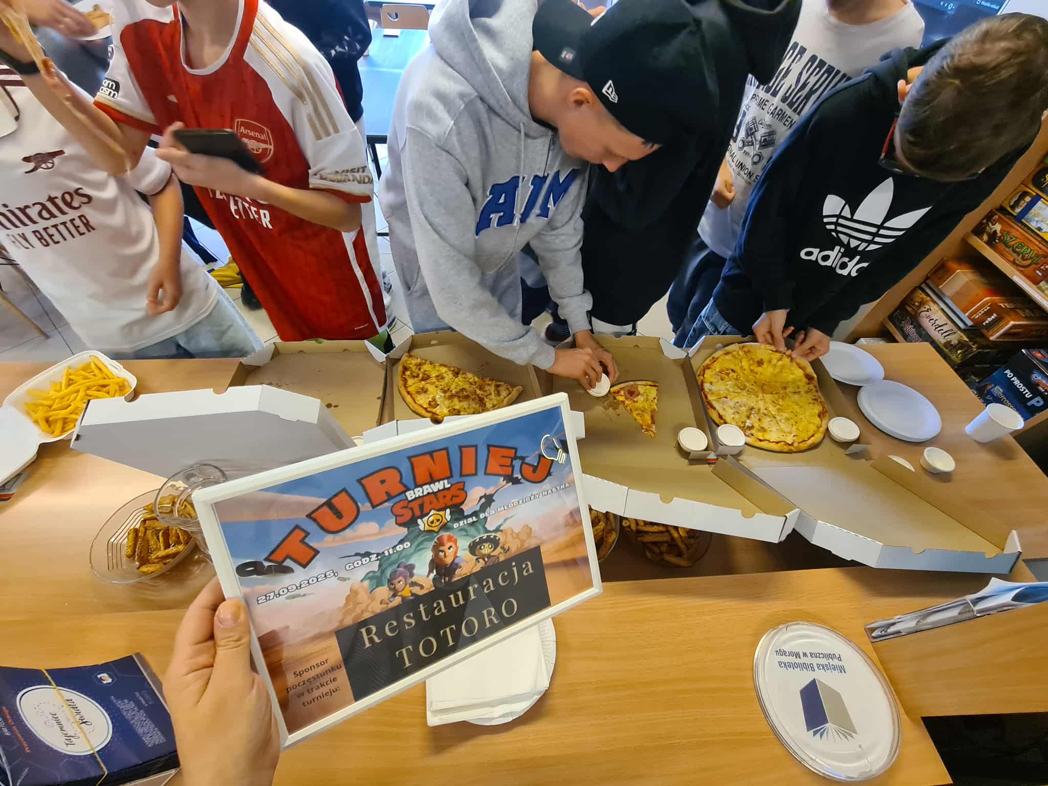 Grupa chłopców kroi i nakłada kawałki pizzy podczas spotkania w bibliotece, na pierwszym planie widoczny plakat turnieju Brawl Stars sponsorowanego przez restaurację Totoro.