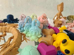Zestaw pastelowych breloczków 3D w kształcie postaci z uszami, obok żółta figurka i inne wydruki.