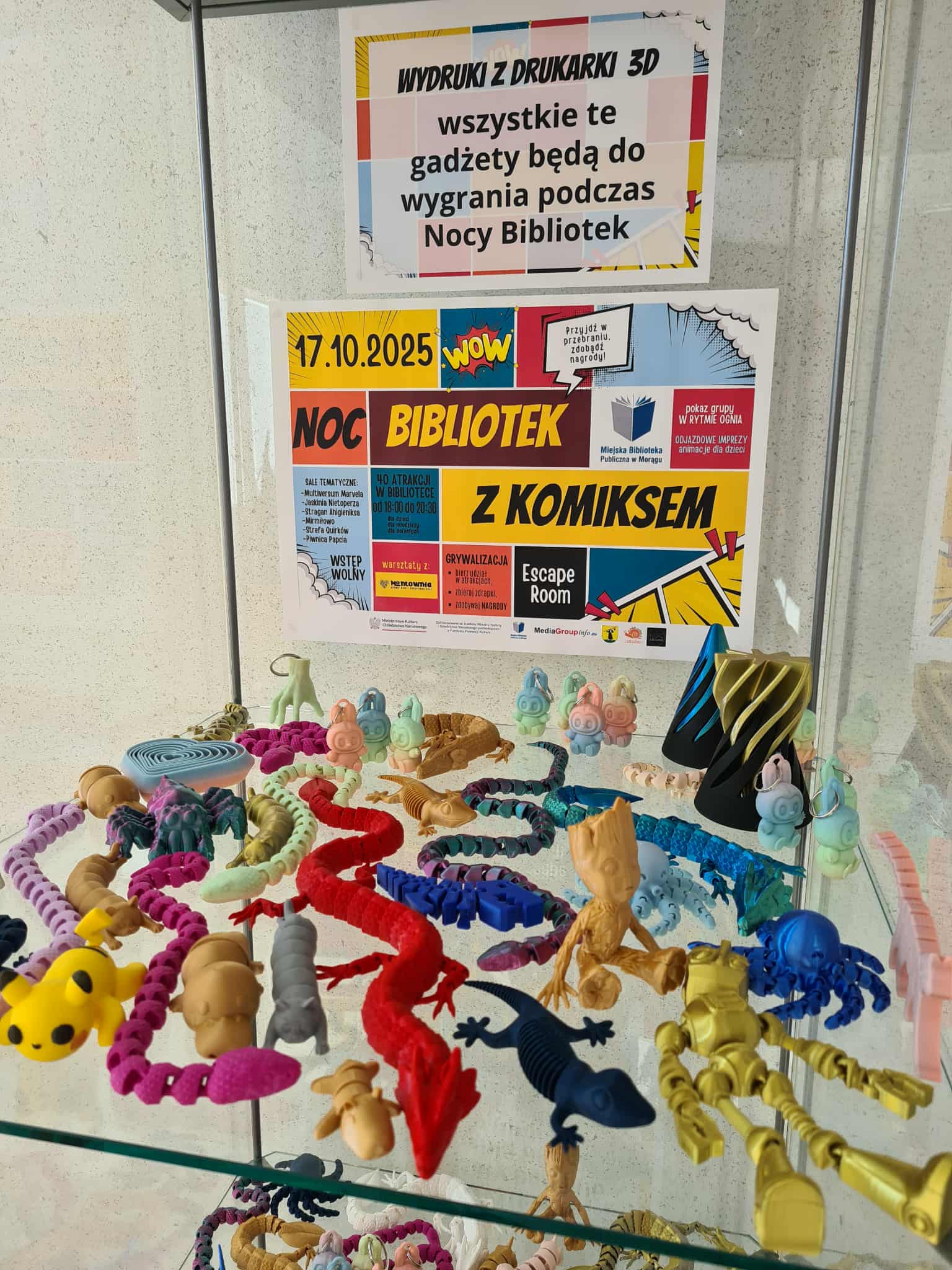 Półka z kolorowymi wydrukami 3D – figurkami zwierząt, postaci i gadżetów, z plakatem zapowiadającym Noc Bibliotek w tle.