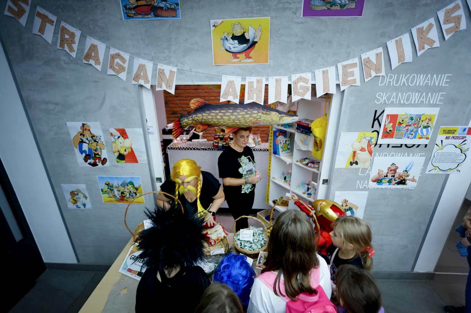 Stoisko „Stragan Ahibieniks” ozdobione ilustracjami z Asteriksa, dzieci kupują gadżety.