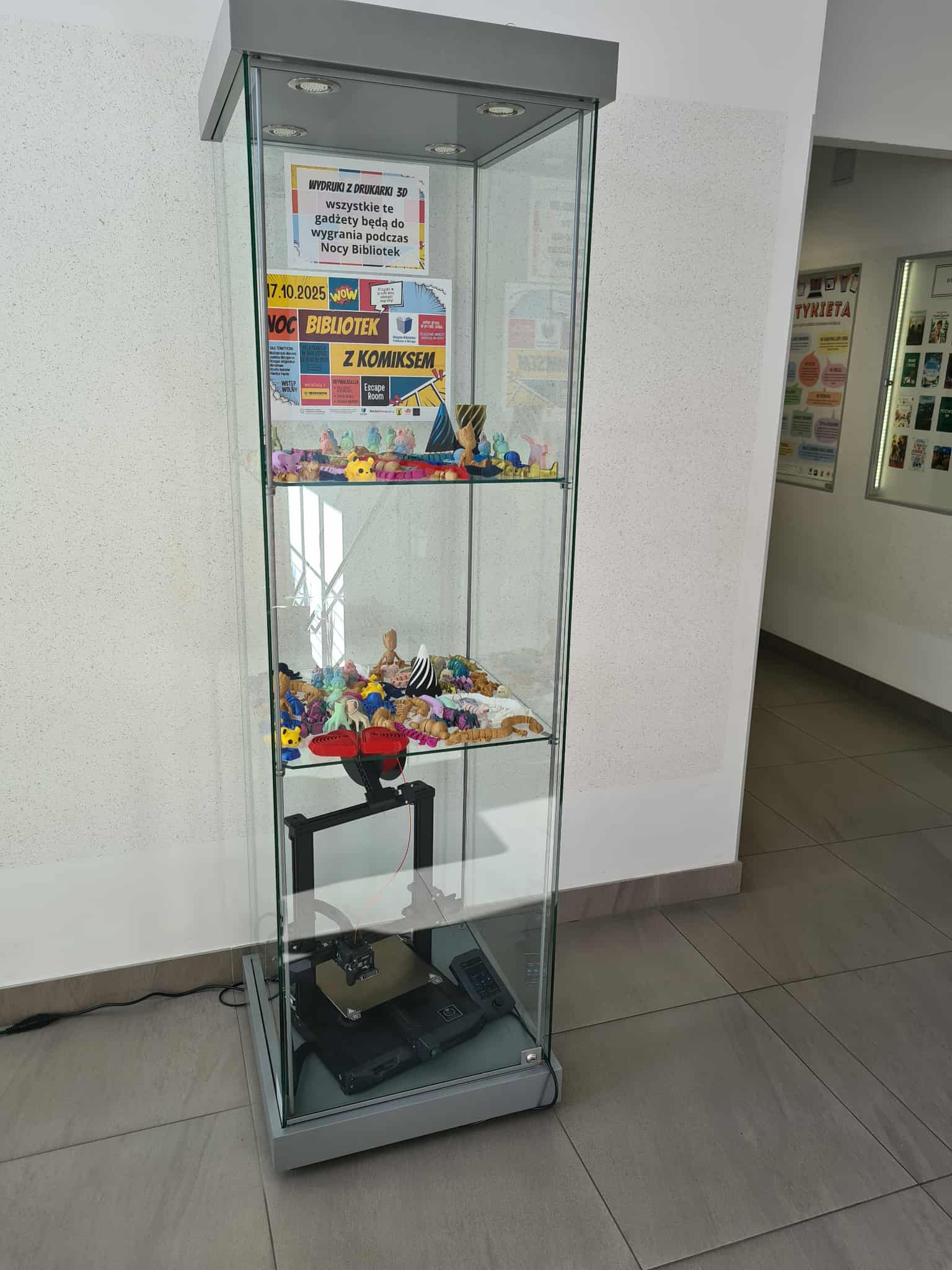 Szklana gablota z drukarką 3D i wystawą wydruków – kolorowe figurki i plakat informujący o Nocy Bibliotek.