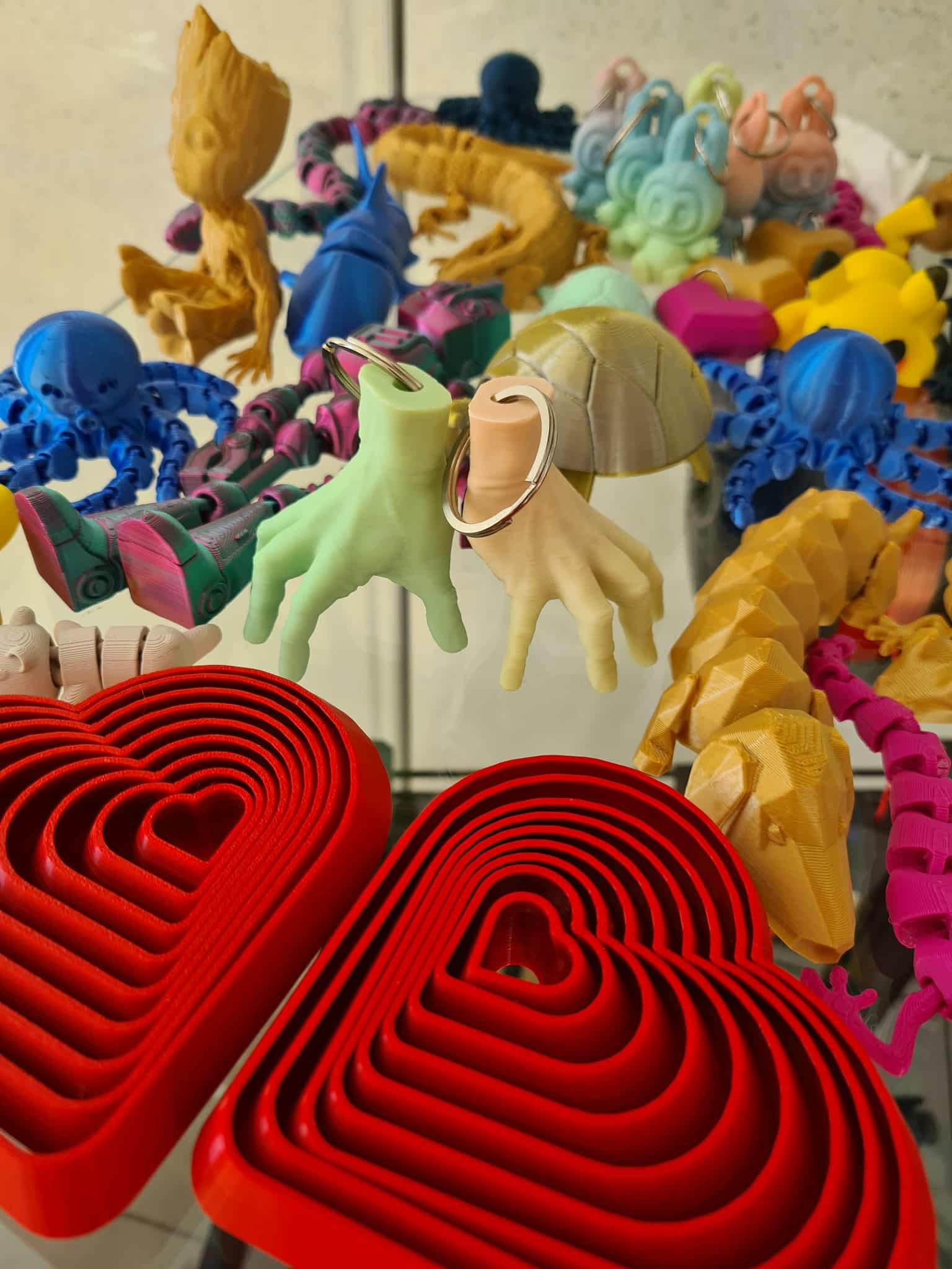 Zbliżenie na wydruki 3D – czerwone serca w kształcie ramek i pastelowe breloczki w kształcie dłoni oraz innych postaci.