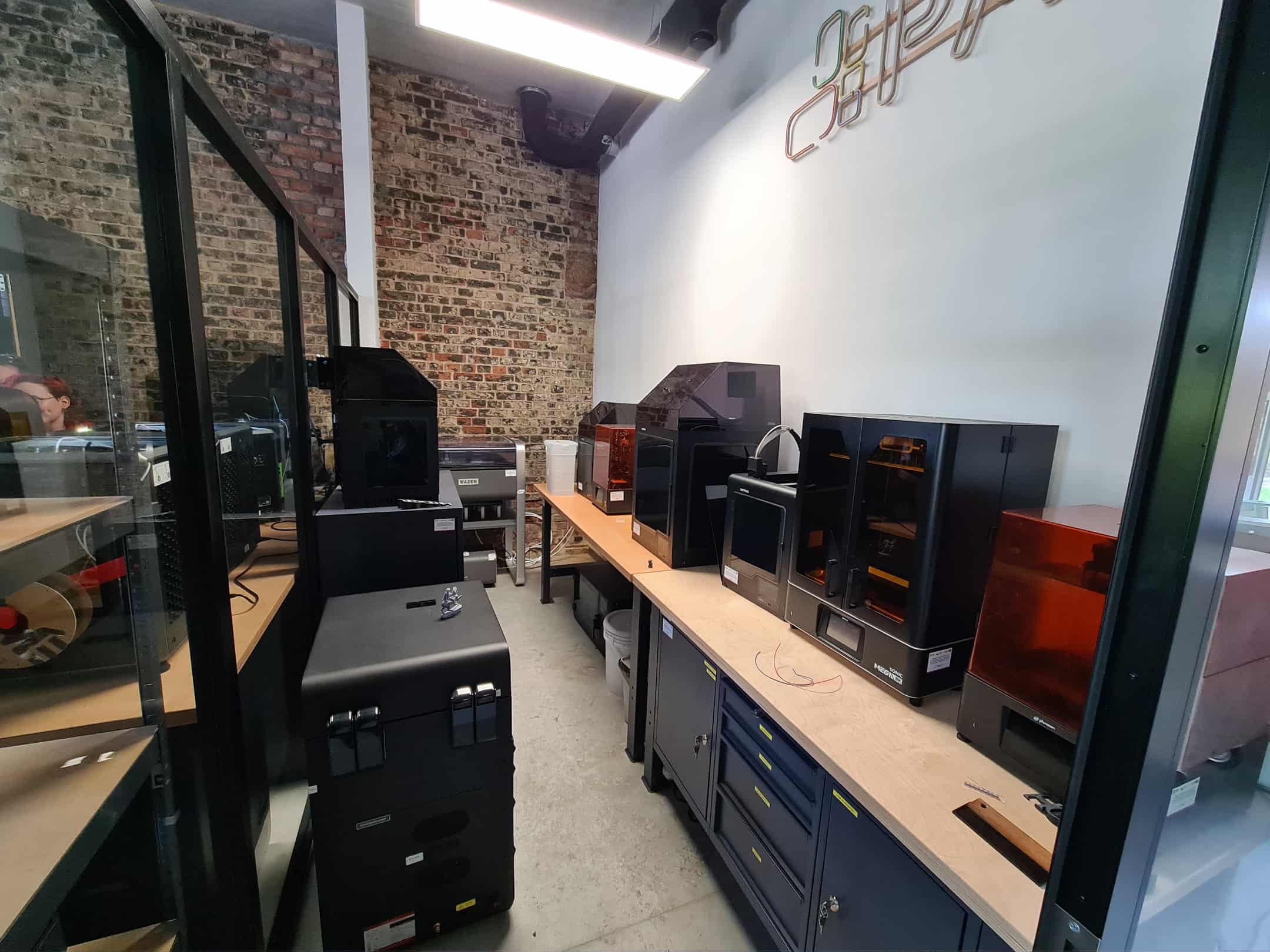 Rząd drukarek 3D ustawionych na stole przy ceglanej ścianie.