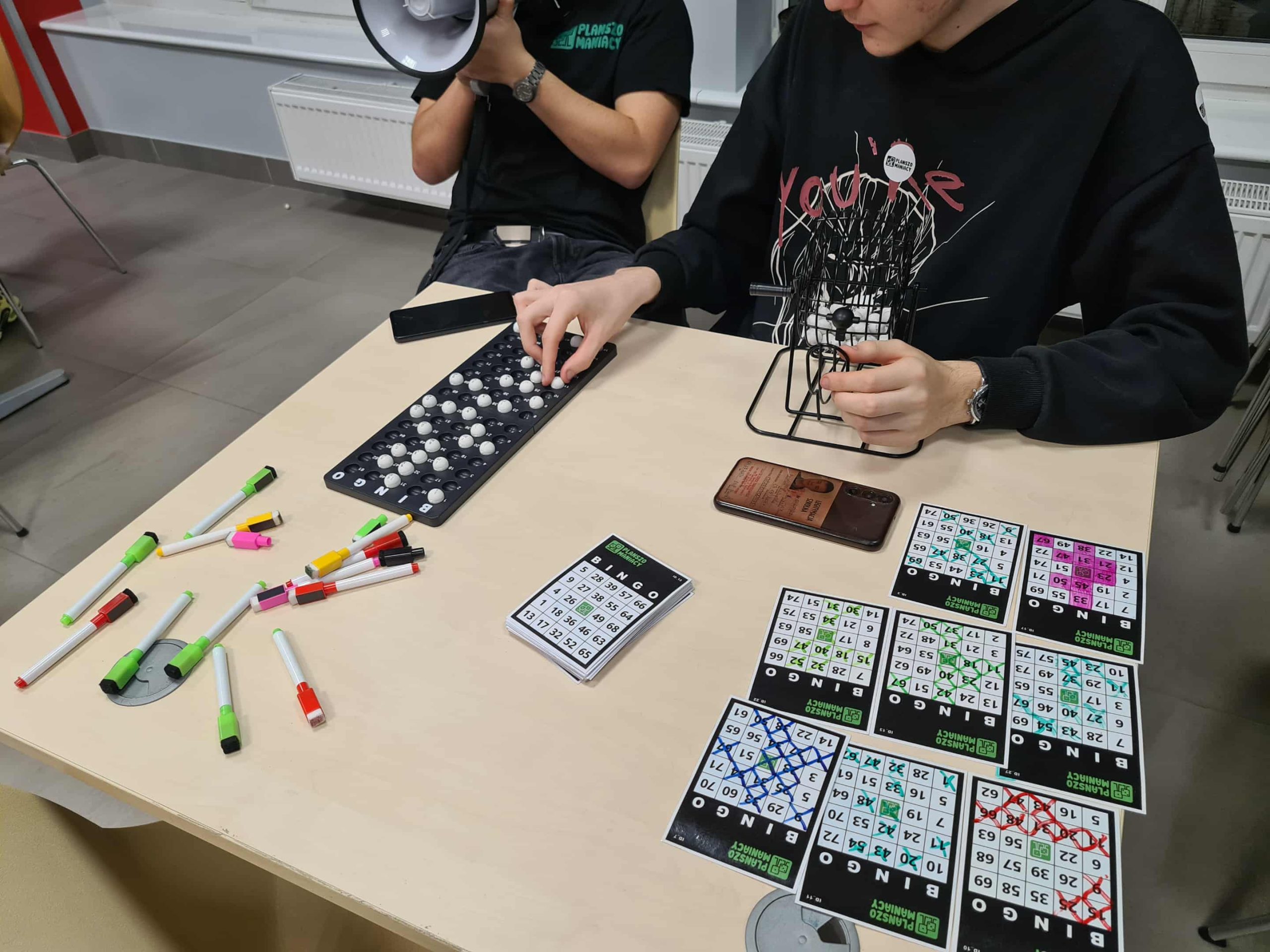 Młodzież gra w bingo, na stole karty, markery i koszyk z kulkami.