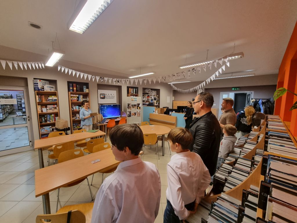 Grupa dorosłych i dzieci stoi w dziale młodzieżowym biblioteki, słuchając osoby prowadzącej spotkanie. W tle widać regały z grami planszowymi i dekoracyjne girlandy.