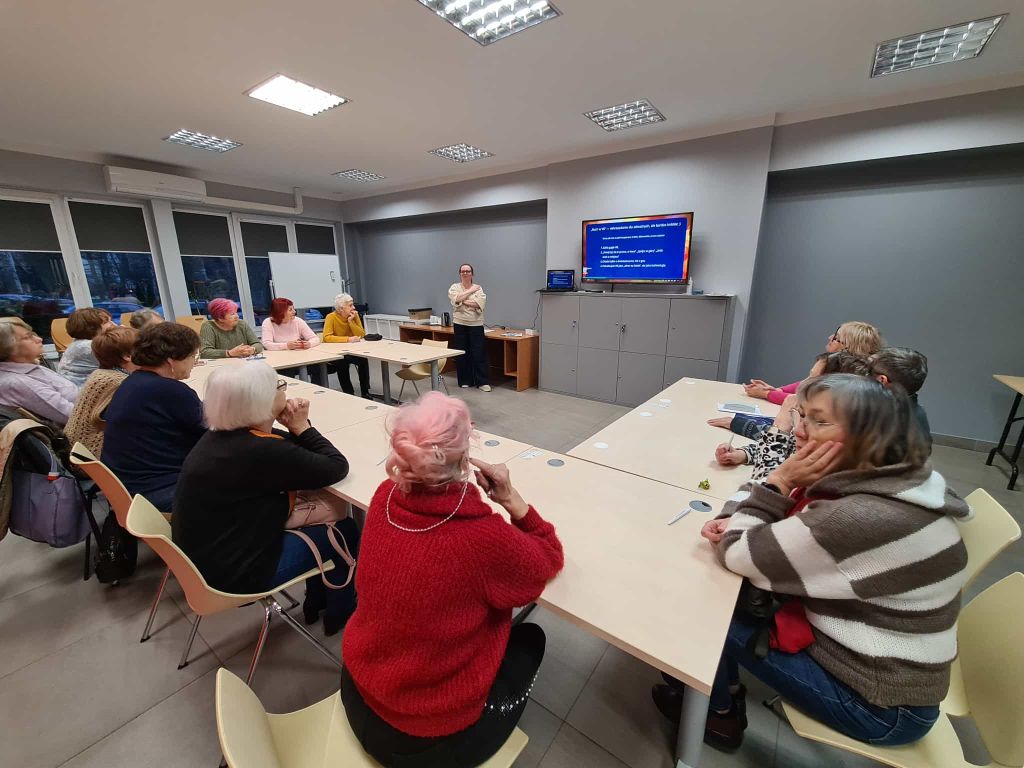 Warsztat w bibliotece – przy dużym stole siedzi grupa seniorów, słuchających osoby prowadzącej stojącej przy ekranie z prezentacją.