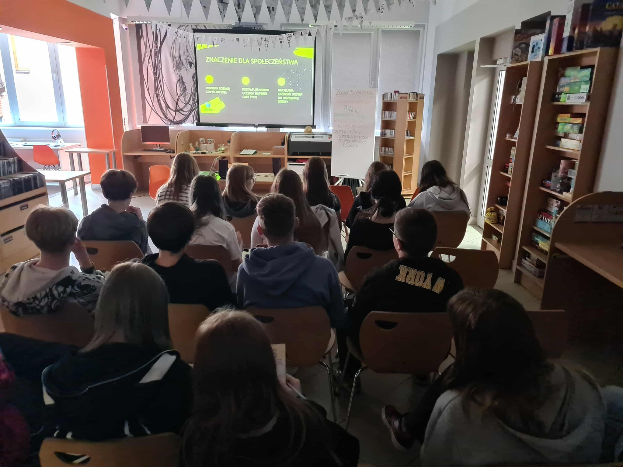 Grupa młodzieży ogląda prezentację o znaczeniu bibliotek dla społeczeństwa na dużym ekranie.