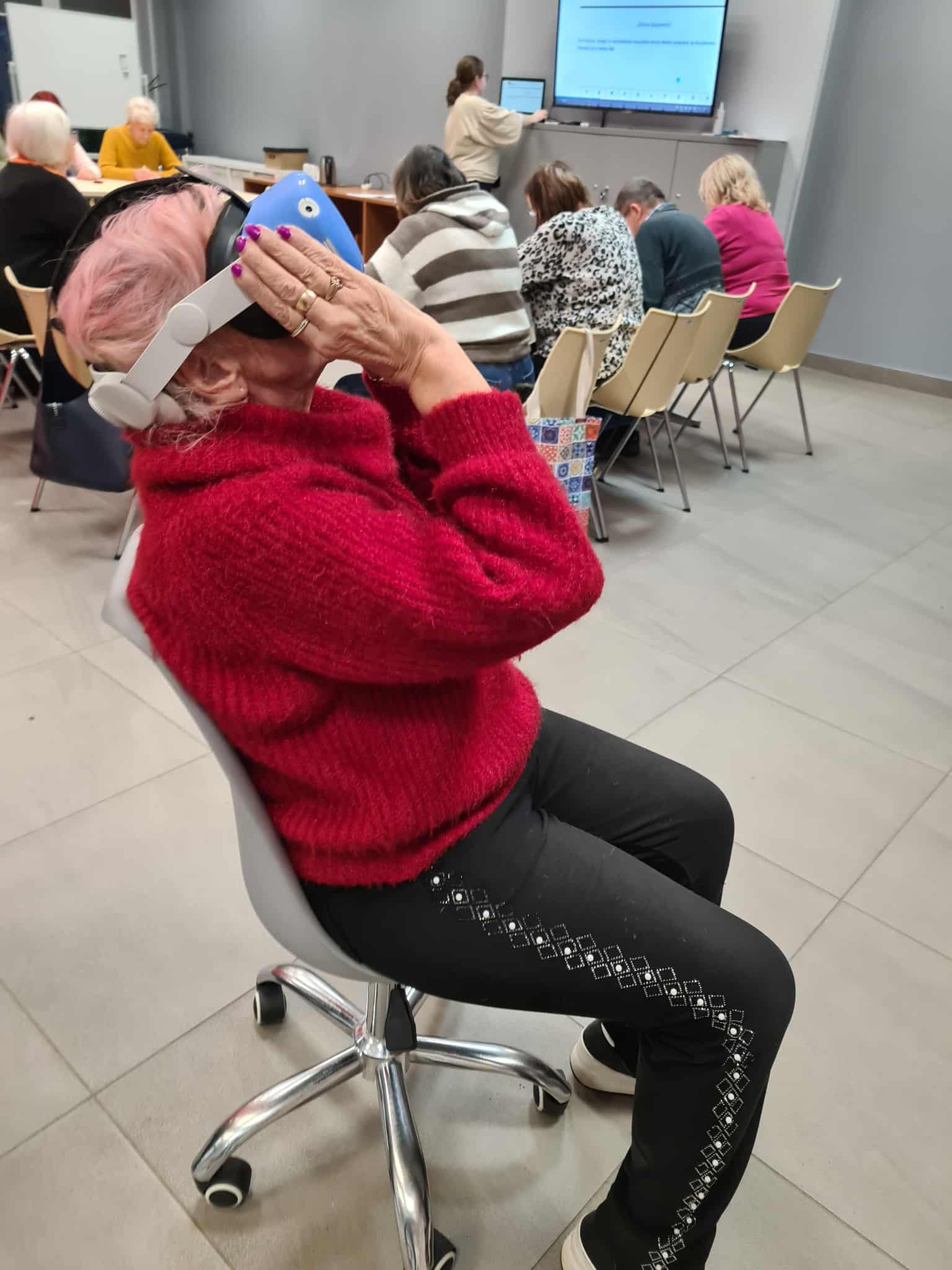 „Starsza kobieta siedzi na krześle w bibliotece i trzyma na głowie gogle VR, patrząc w górę. W tle inni uczestnicy zajęć piszą lub słuchają prowadzącej.”