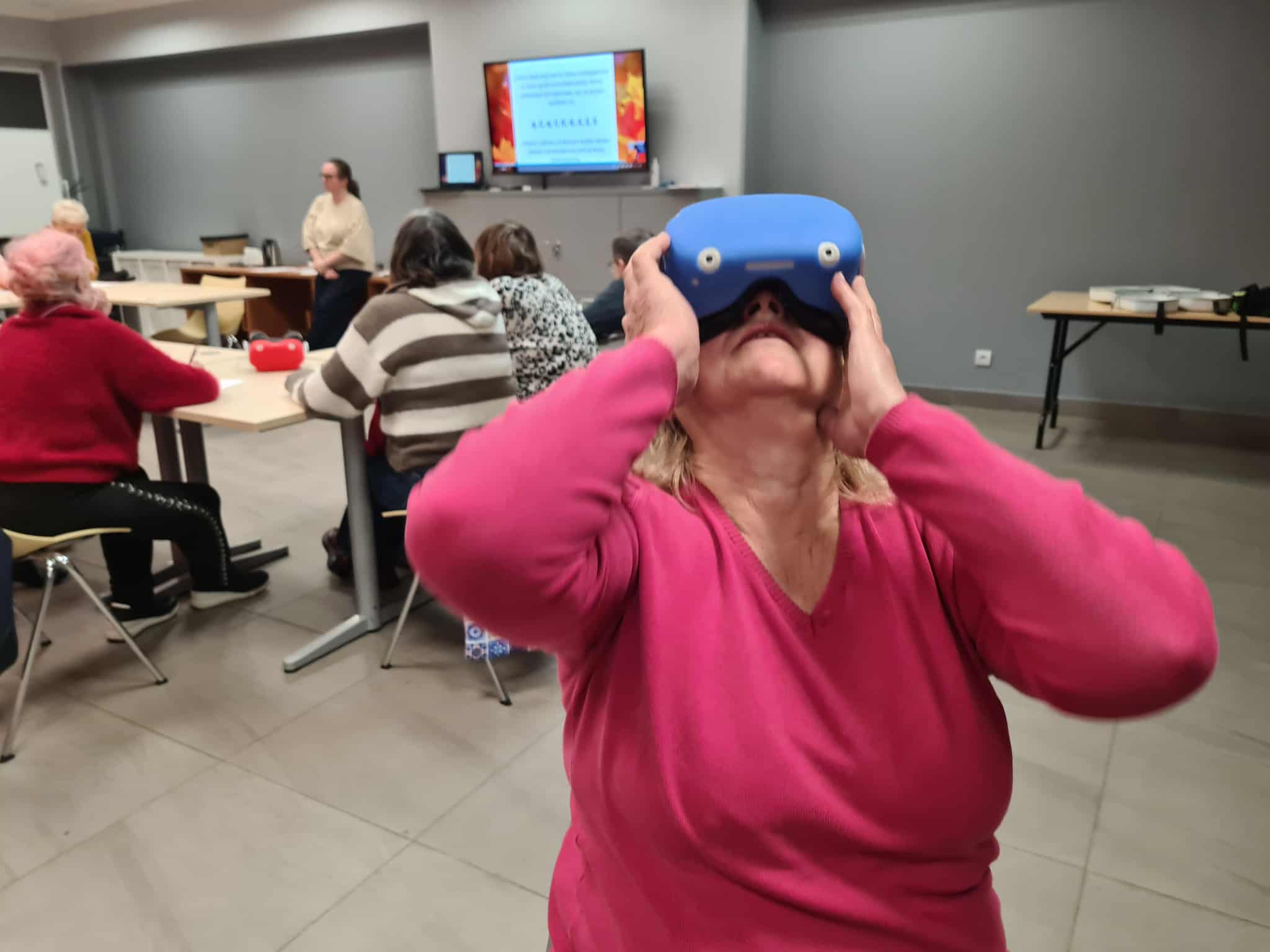 „Kobieta w różowym swetrze trzyma na głowie niebieskie gogle VR podczas zajęć w bibliotece. Za nią pozostali uczestnicy siedzą przy stołach, a prowadząca stoi przy monitorze.”
