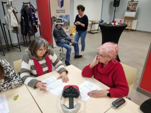 Dwie seniorki pracują przy stoliku nad kartkami z zadaniami, a w tle mężczyzna w okularach VR siedzi na krześle, podczas gdy animatorka mu pomaga.