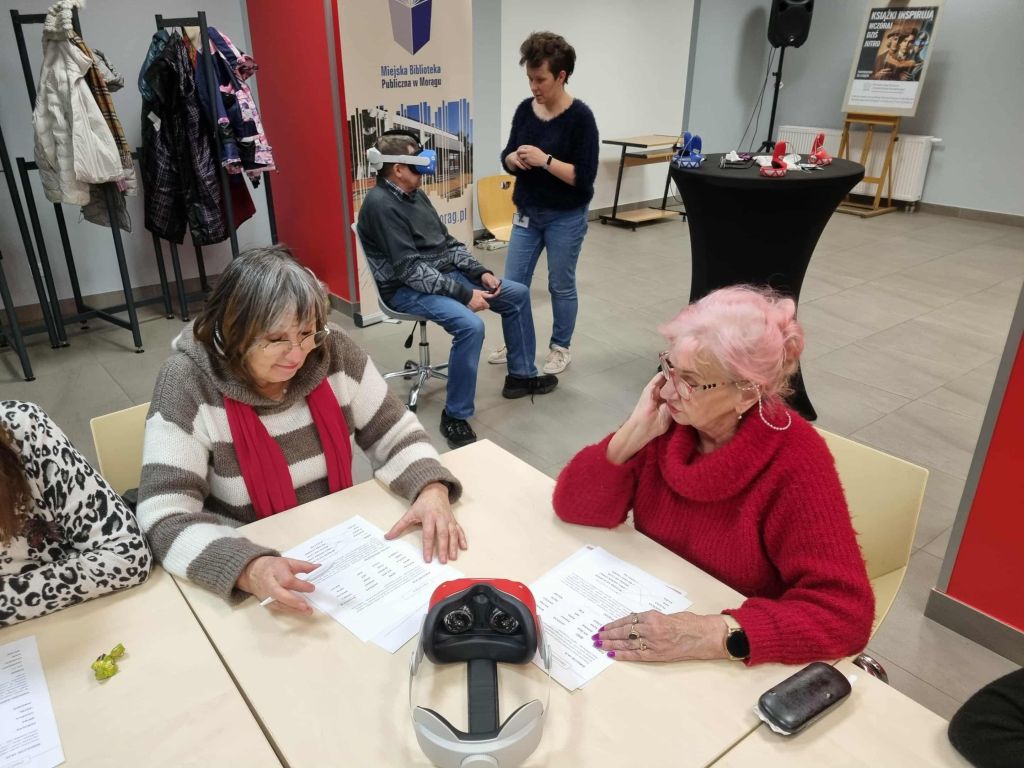 Dwie seniorki pracują przy stoliku nad kartkami z zadaniami, a w tle mężczyzna w okularach VR siedzi na krześle, podczas gdy animatorka mu pomaga.