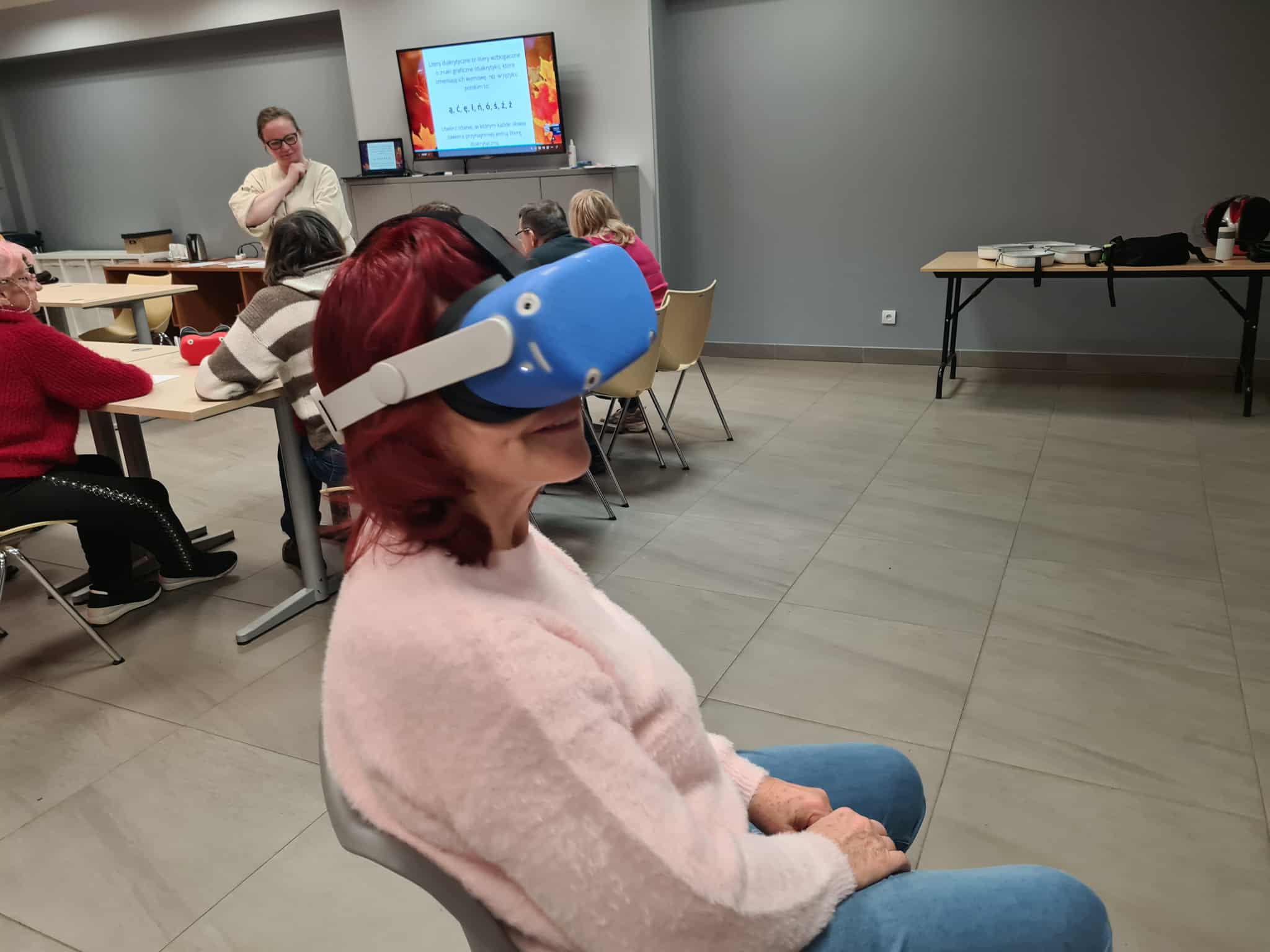 „Uczestniczka warsztatów w bibliotece, ubrana w różowy sweter, siedzi na krześle i ogląda treści w goglach VR. W tle widać osoby pracujące przy stołach oraz prezentację na ekranie.”