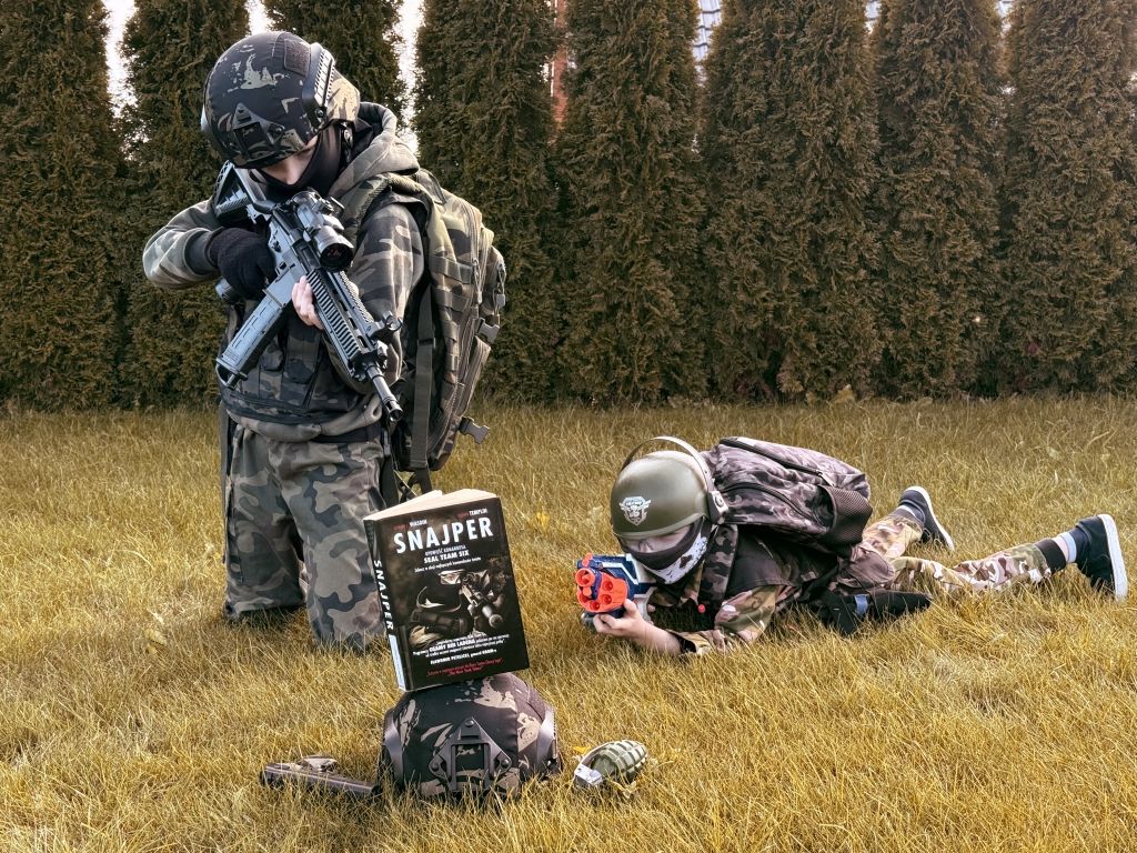 Dwaj chłopcy przebrani w wojskowe stroje pozują na trawie; jeden celuje z zabawkowej broni, drugi leży z plastikowym pistoletem. Przed nimi stoi książka „Snajper”.