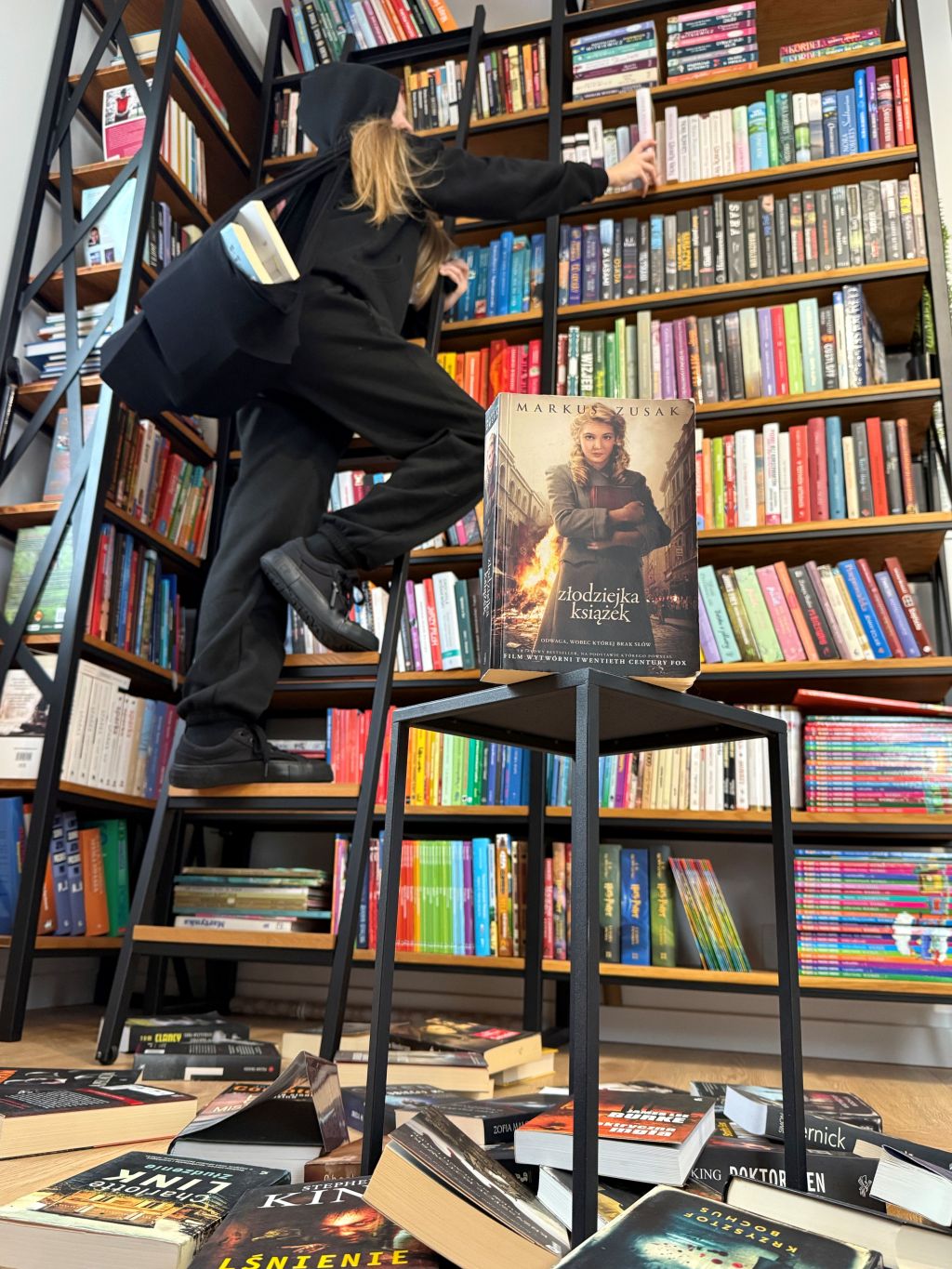 Dziewczyna ubrana na czarno wspina się po drabinie przy regale wypełnionym książkami, z tomem wystającym z torby. Na pierwszym planie stoi książka „Złodziejka książek”, a na podłodze leżą porozrzucane powieści.