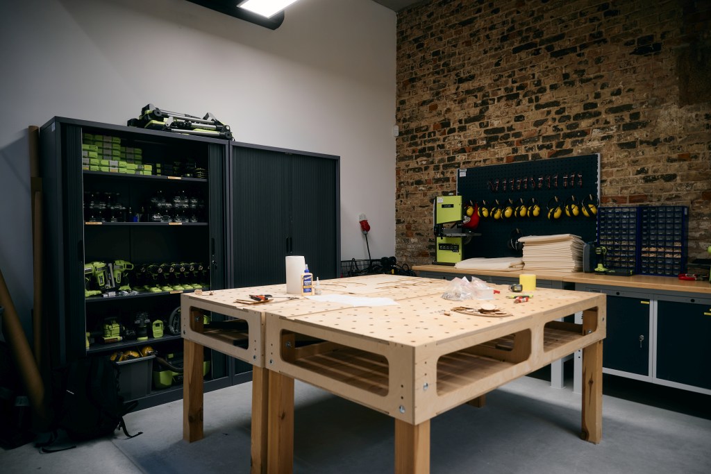 Warsztat z dużym stołem roboczym, narzędziami i sprzętem stolarskim na tle ceglanej ściany.