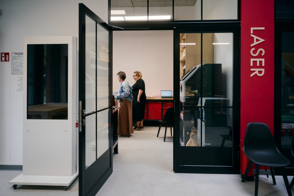 Widok przez przeszklone drzwi do pomieszczenia z laserem, w środku dwie osoby przy komputerze.