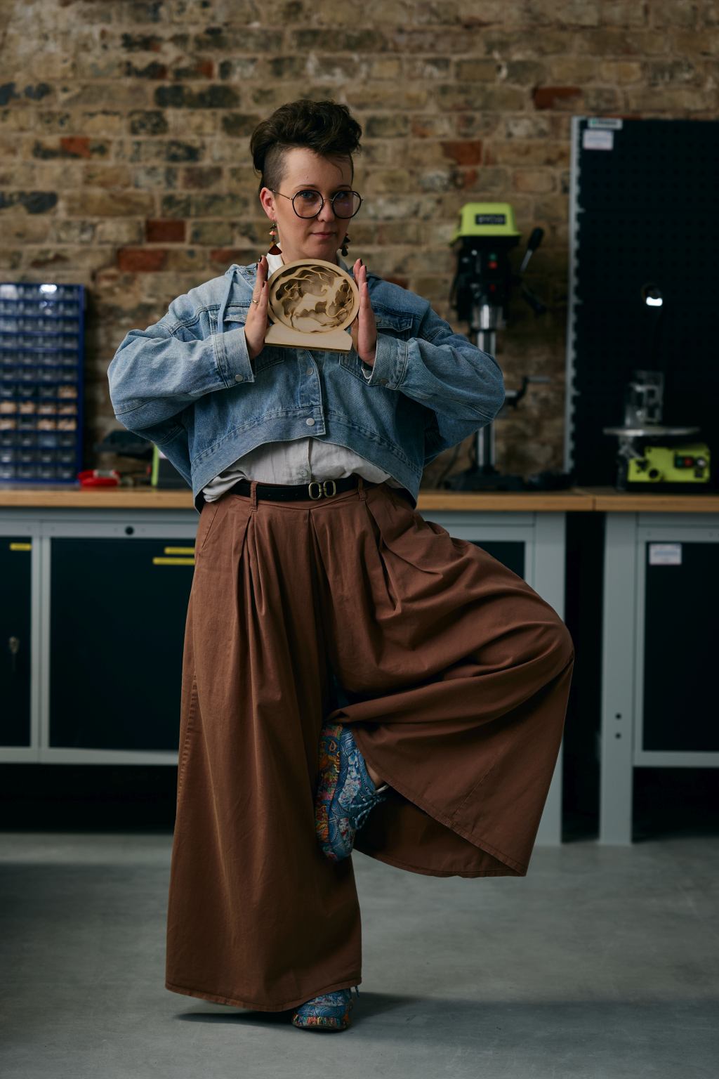 Osoba w jeansowej kurtce i szerokich spodniach prezentuje drewniany wyrób z laserowej wycinarki.