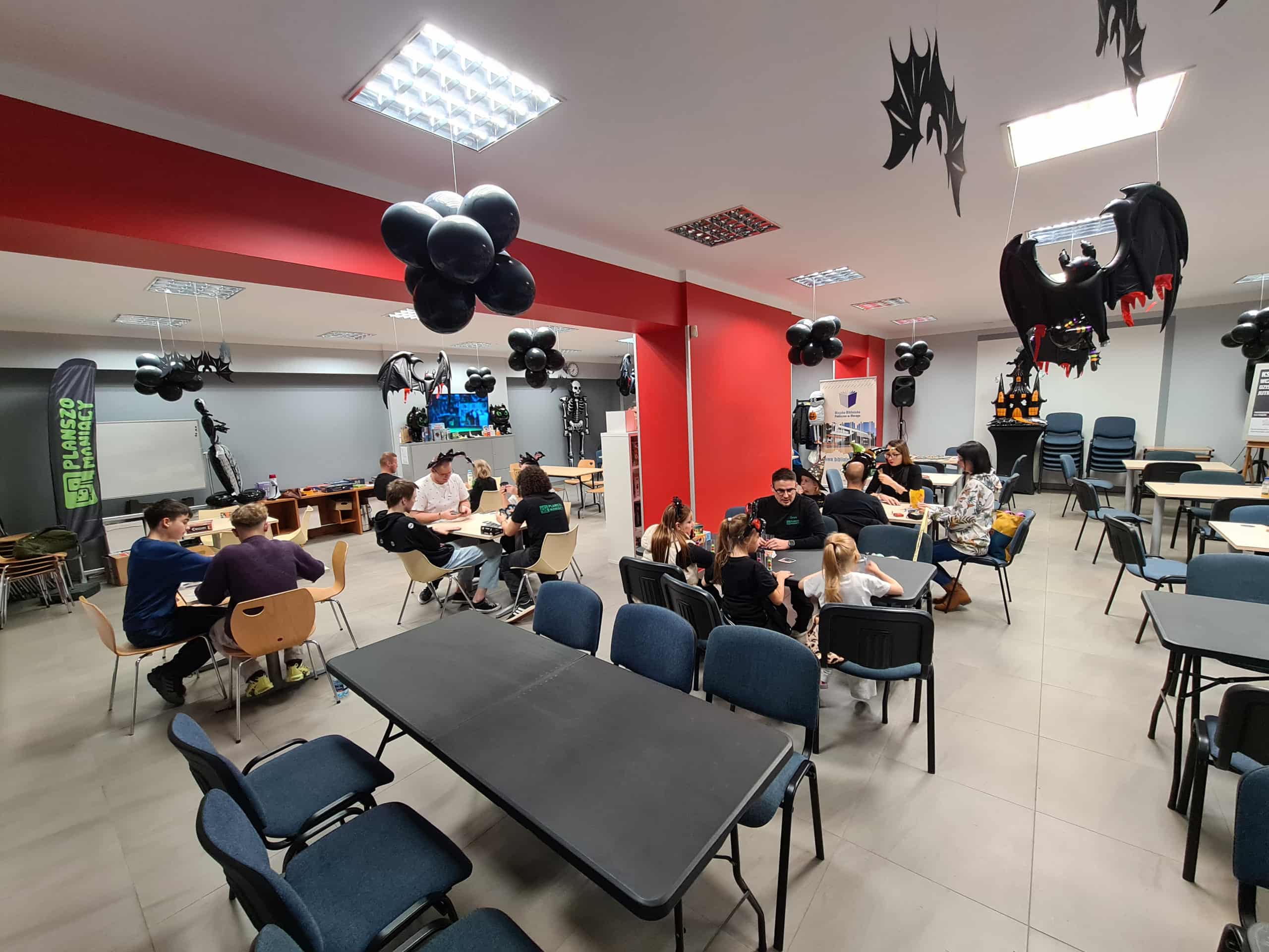 Sala biblioteczna udekorowana czarnymi balonami i smokami, uczestnicy grają przy stołach.