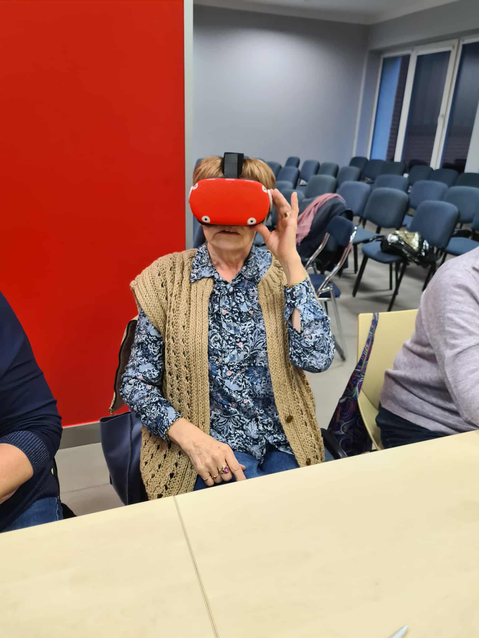 Seniorka siedzi przy stole i trzyma na głowie czerwone gogle VR, testując wirtualną rzeczywistość.