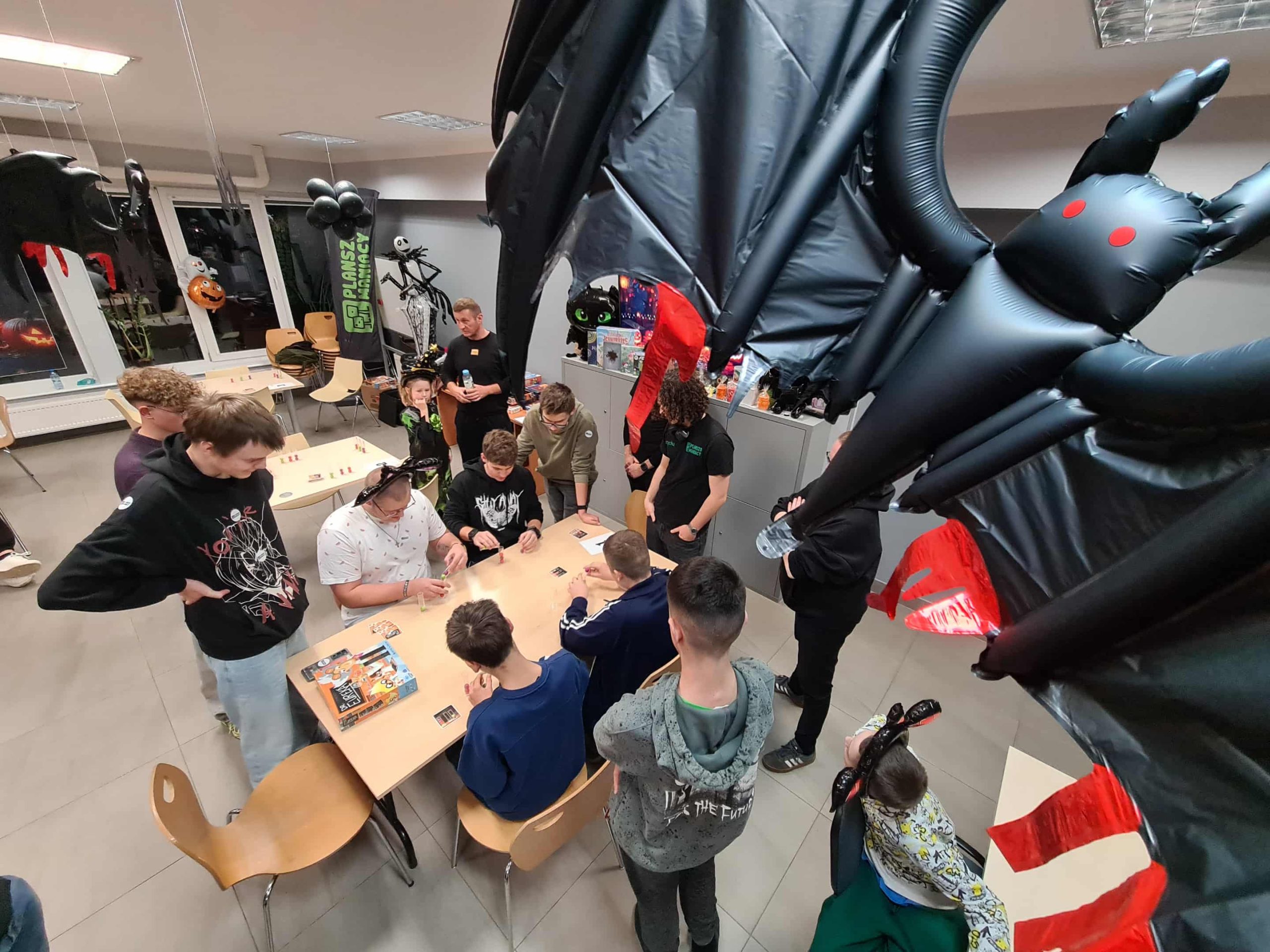 Młodzież i prowadzący grają w gry planszowe przy stole, nad nimi zawieszony czarny smok.