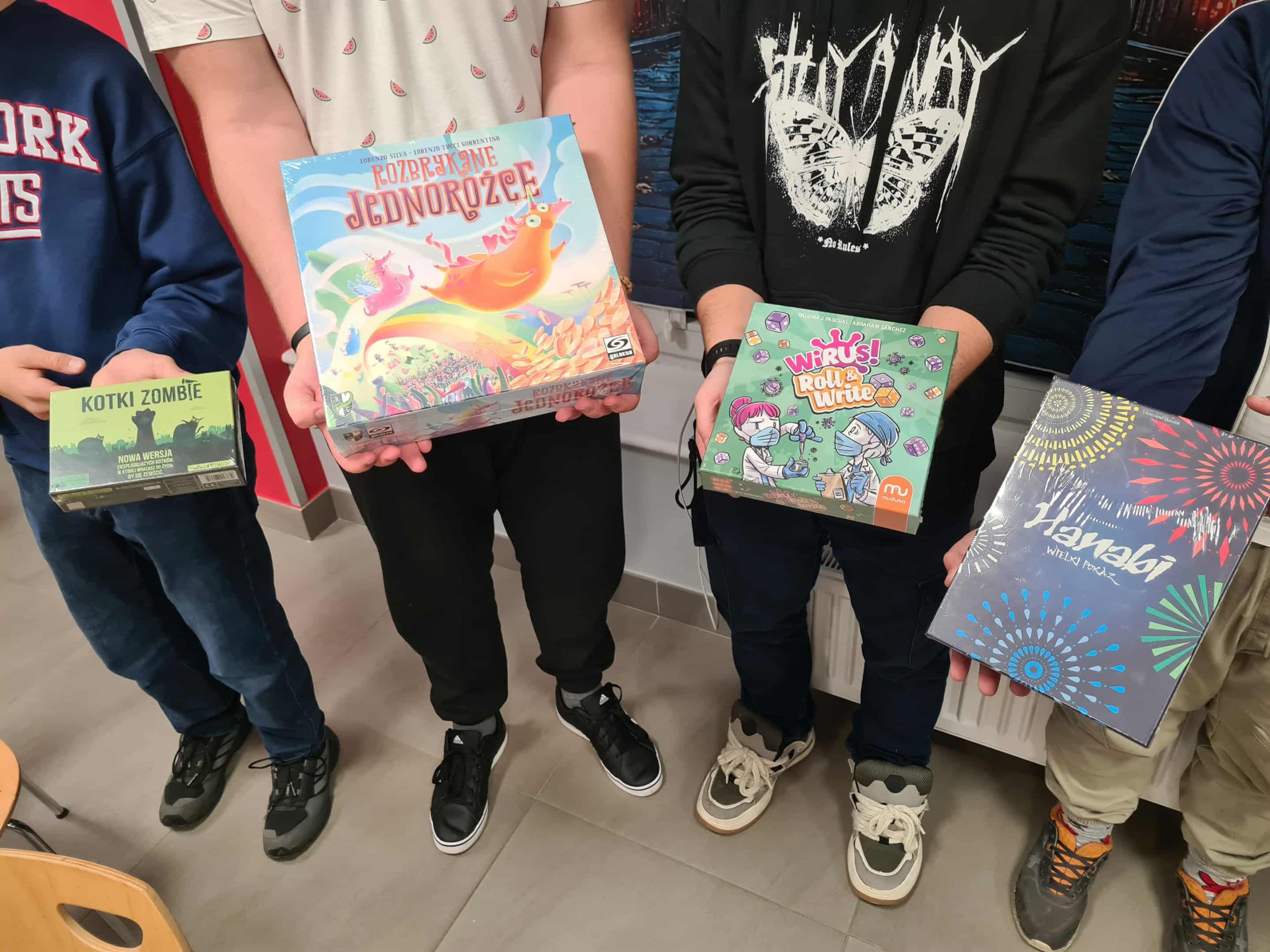 Uczestnicy pokazują pudełka z grami planszowymi: „Kotki Zombie”, „Rozbrykane Jednorożce”, „Wirus!” i „Hanabi”.