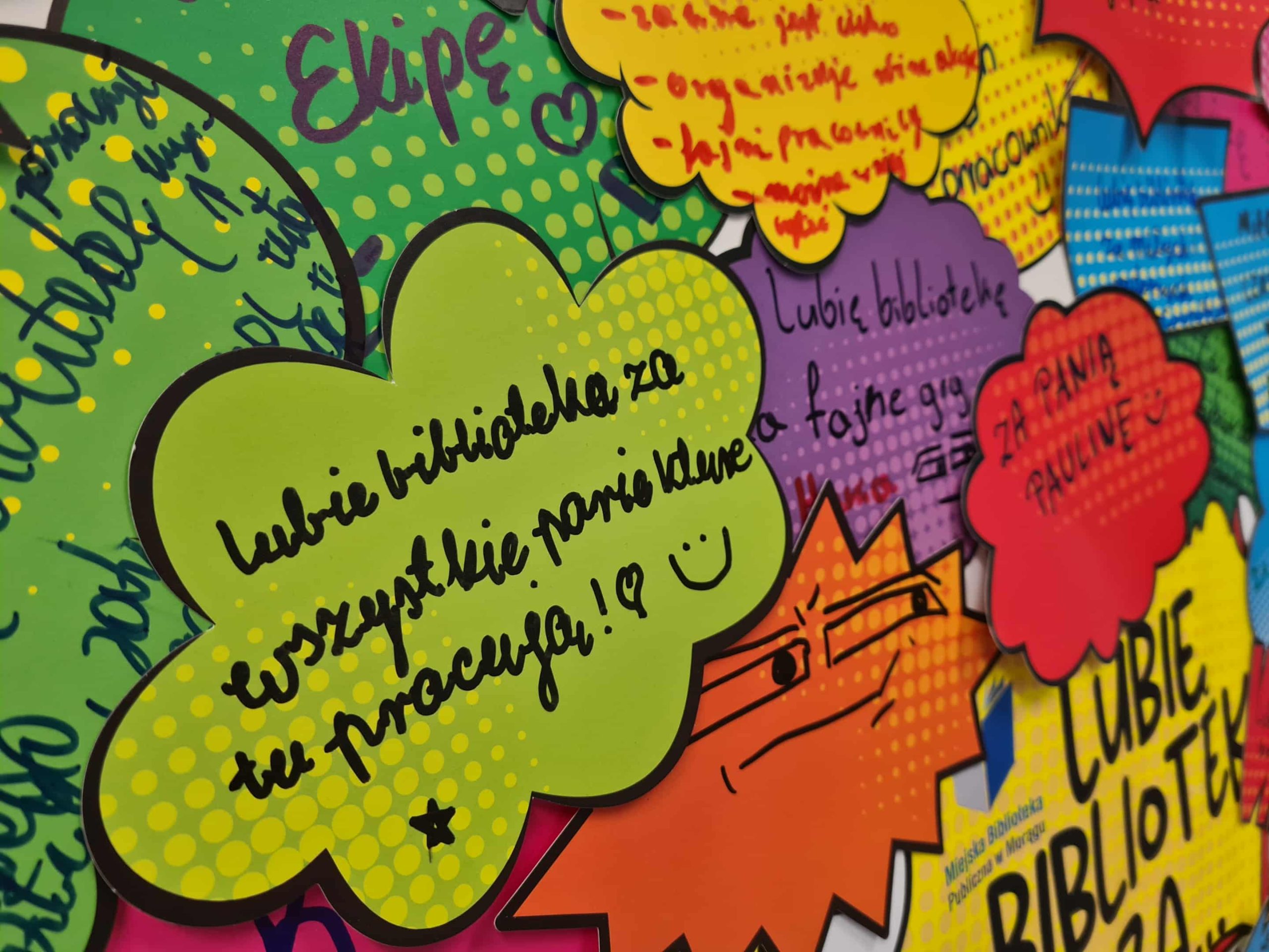 Zielona chmurka z napisem „Lubię bibliotekę za wszystkie panie, które tu pracują” oraz kolorowe dymki wokół.